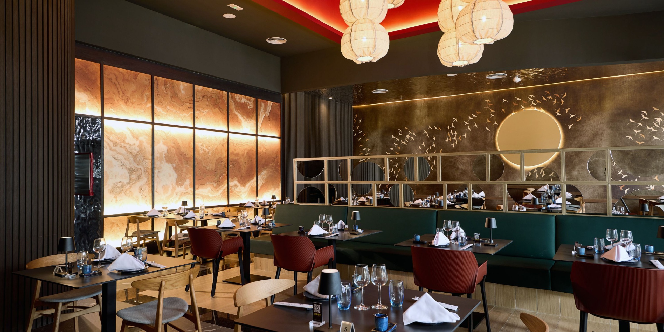 Modern dining setup at Hotel Riu Palace Bavaro, showcasing tables ready for guests, wooden chairs with red cushioned seats, ambient lighting, and decorative wall art. Ceiling is adorned with large hanging lamps.