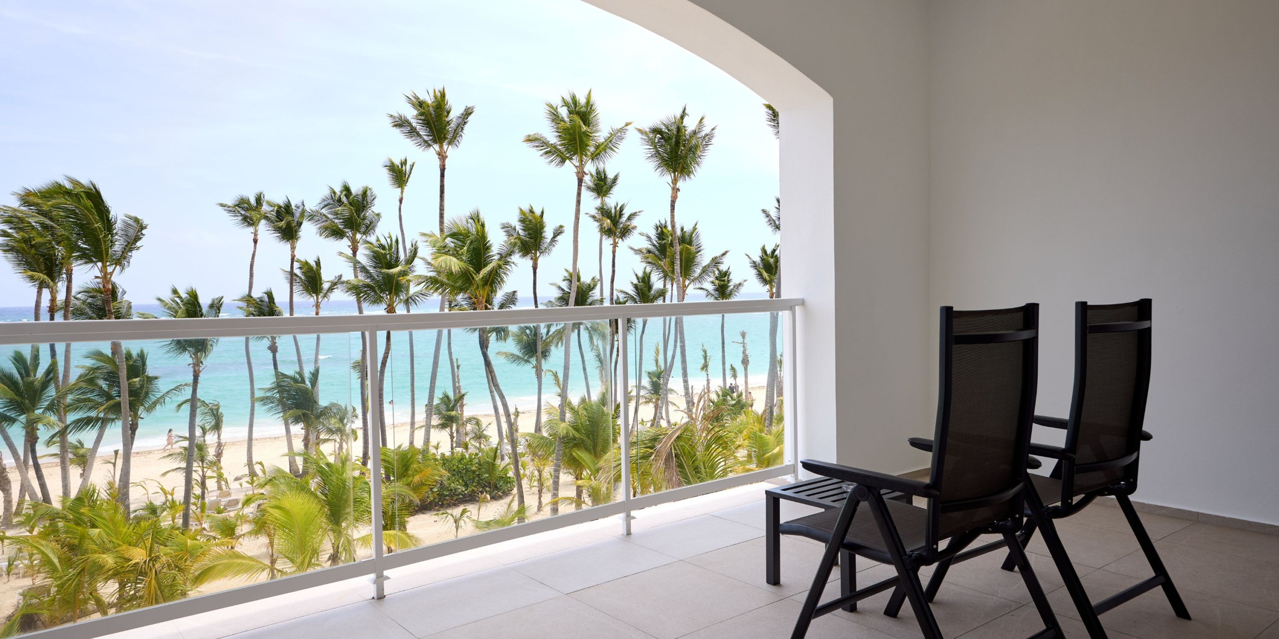 A balcony at Hotel Riu Palace Bavaro features two chairs with a view of the beach, lush palm trees, and the ocean stretching into the distance.