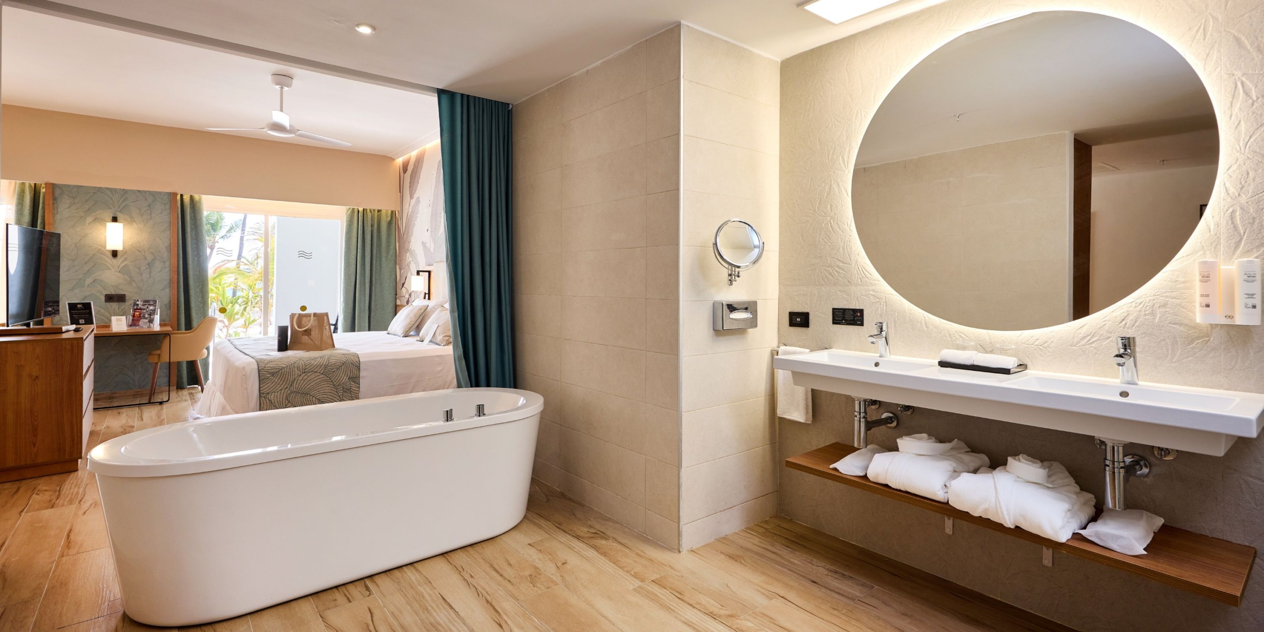 The Hotel Riu Palace Bavaro offers a modernly designed room that includes a bathtub, a double sink vanity, and a view into the bedroom featuring a bed, TV, and access to the balcony.