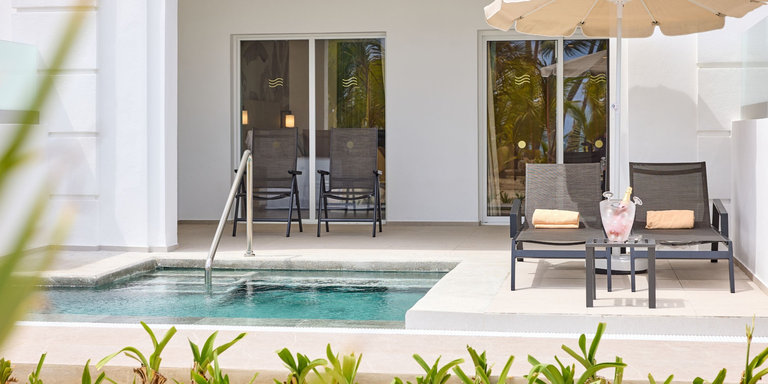 The scene shows a patio at Hotel Riu Palace Bavaro featuring two lounge chairs and a table beside a small pool. Glass doors and additional chairs can be seen in the background, with an umbrella partially visible above the chairs.