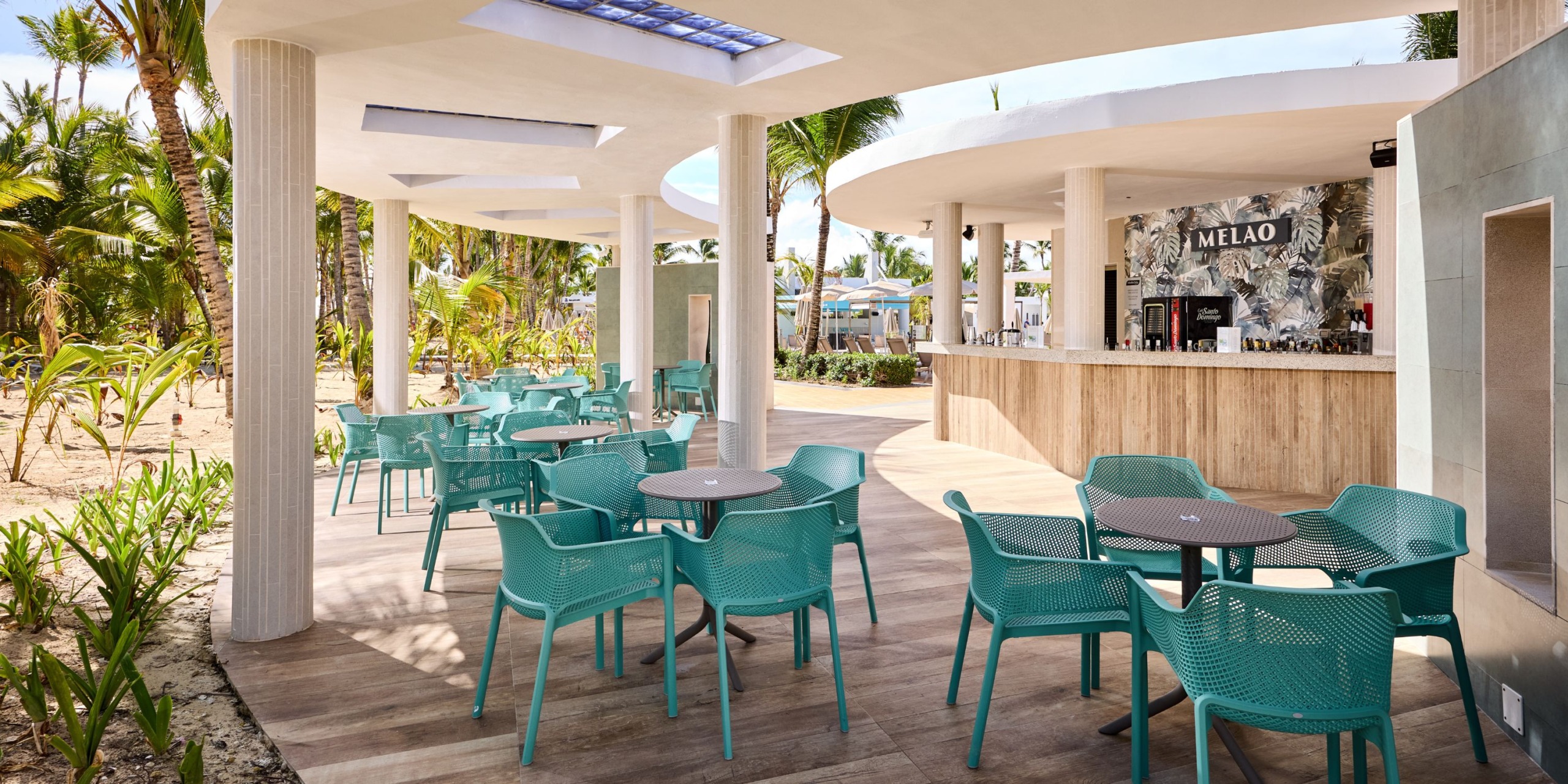 An outdoor cafe with turquoise chairs and round tables is nestled among palm trees at the Hotel Riu Palace Bavaro, with a bar featuring a "Melao" sign in the background.