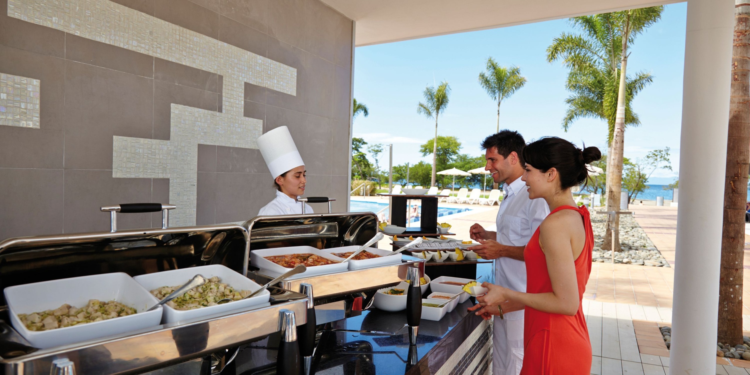 A chef in a white uniform serves a man and woman at an outdoor buffet with various dishes at the Hotel Riu Palace Costa Rica, where palm trees and a pool can be seen in the background.