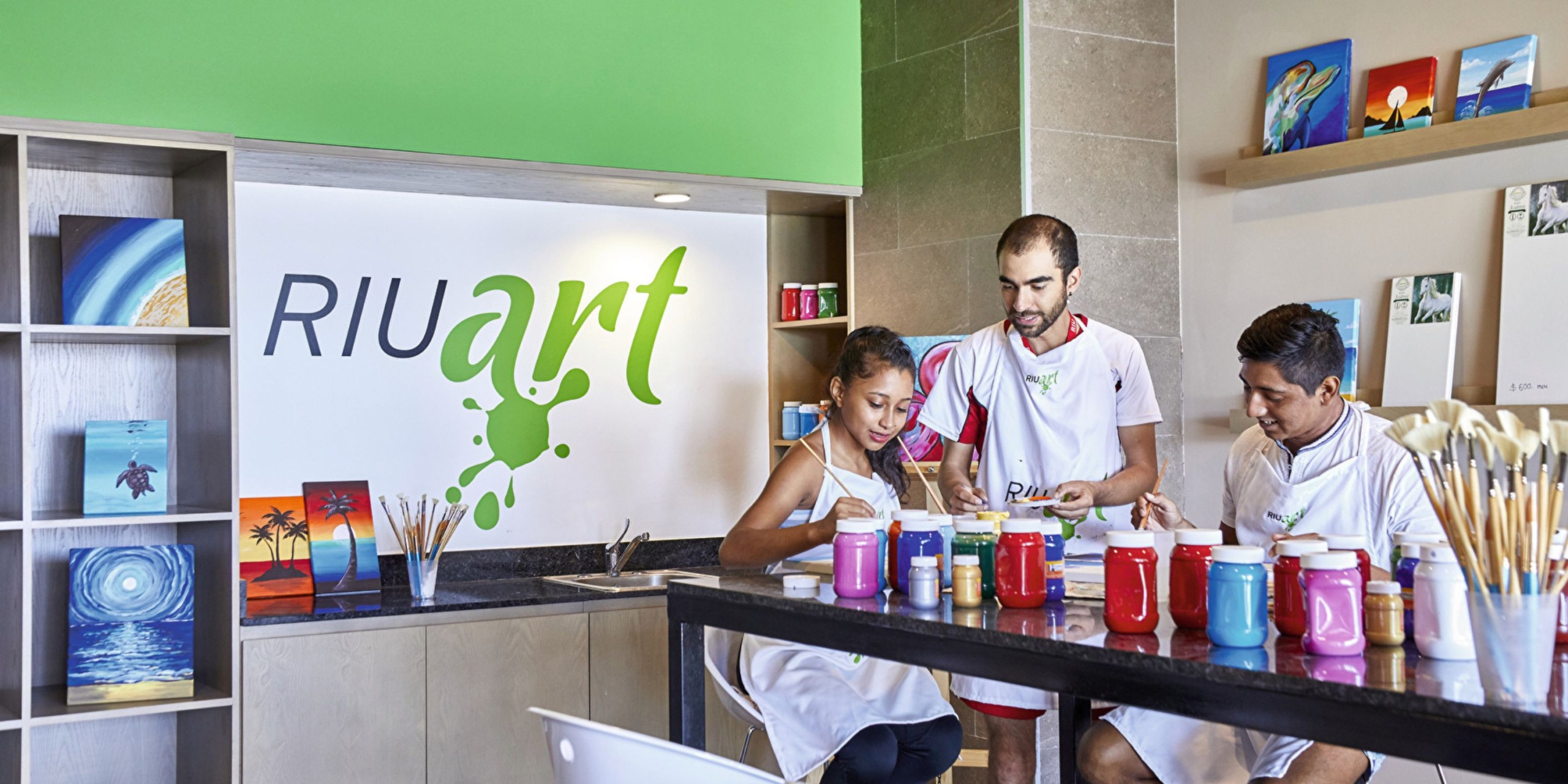 Three individuals are painting at a table filled with various colored jars and brushes, inside a room labeled "Hotel Riu Palace Costa Mujeres Art" with paintings displayed on shelves.