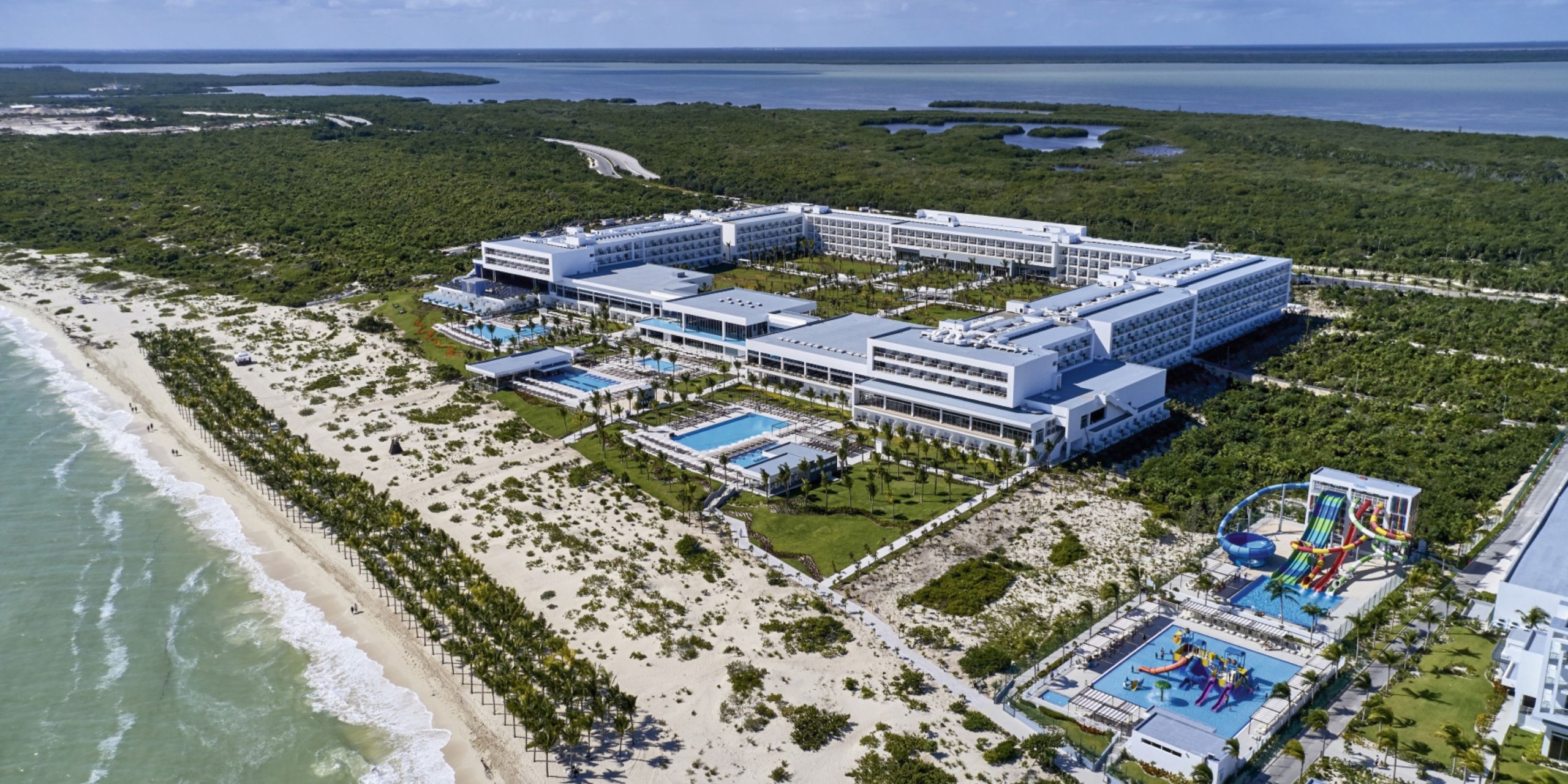 Aerial view of the expansive Hotel Riu Palace Costa Mujeres, featuring multiple pools and a waterpark, set amidst lush greenery and overlooking the vast ocean.