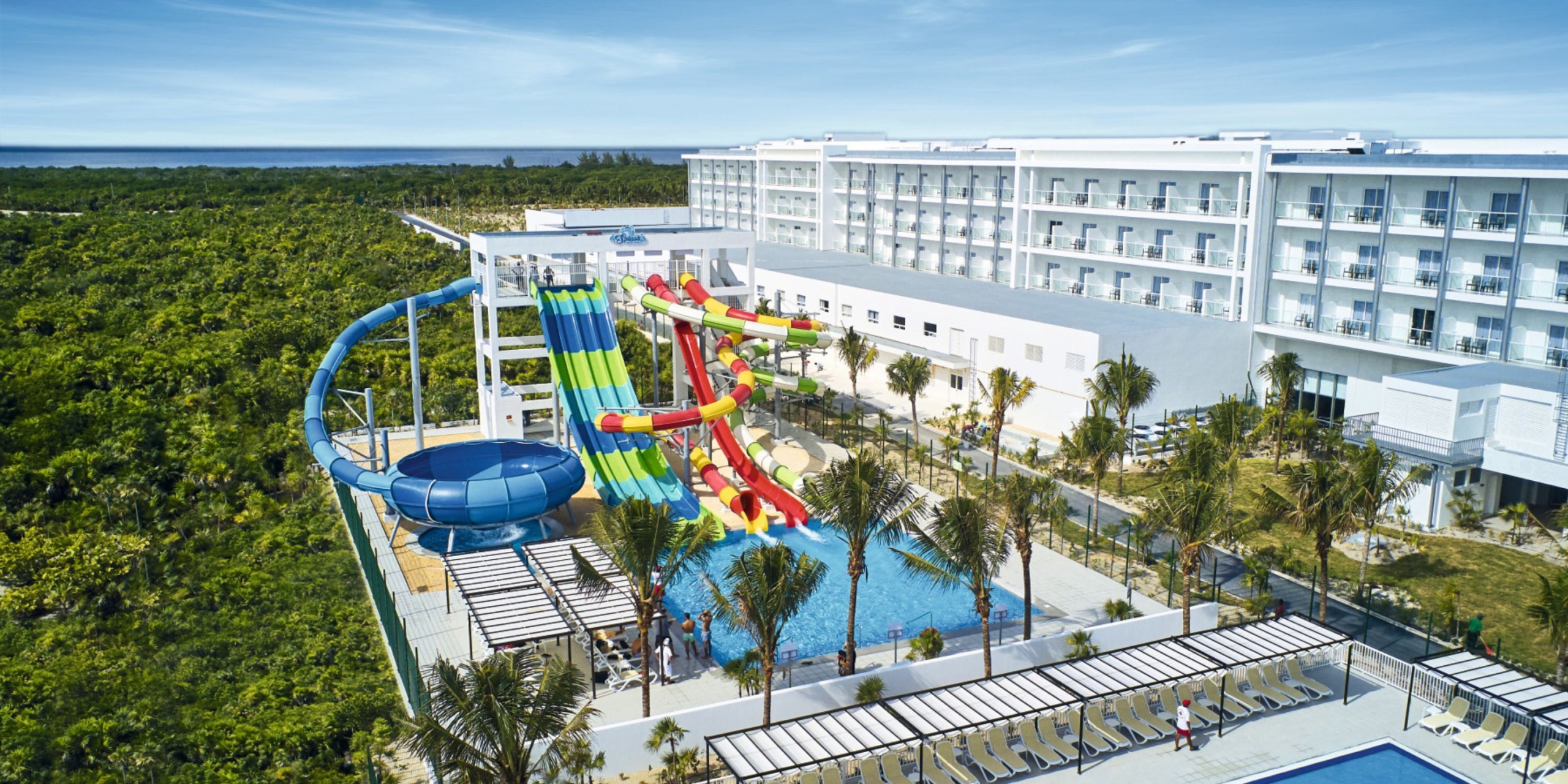 Aerial view of Hotel Riu Palace Costa Mujeres showcasing its sleek white structure, a sparkling pool, vibrant water slides, and lush palm trees set against a backdrop of clear blue skies.