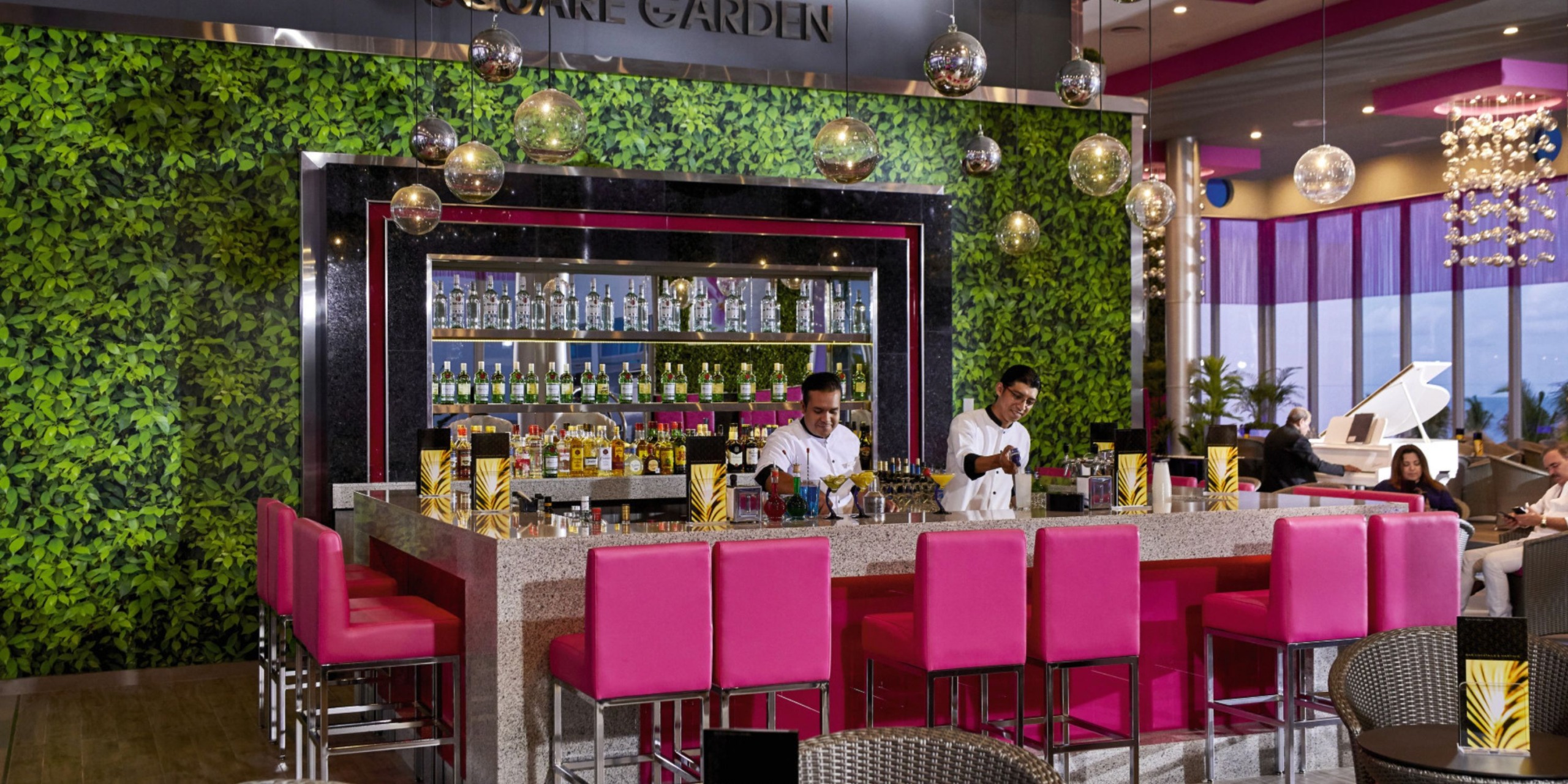 The scene at Hotel Riu Palace Costa Mujeres includes a chic bar featuring two bartenders, with pink chairs and a vibrant green plant wall. In the background, shelves display an array of bottles, while round pendant lights elegantly hang from the ceiling.