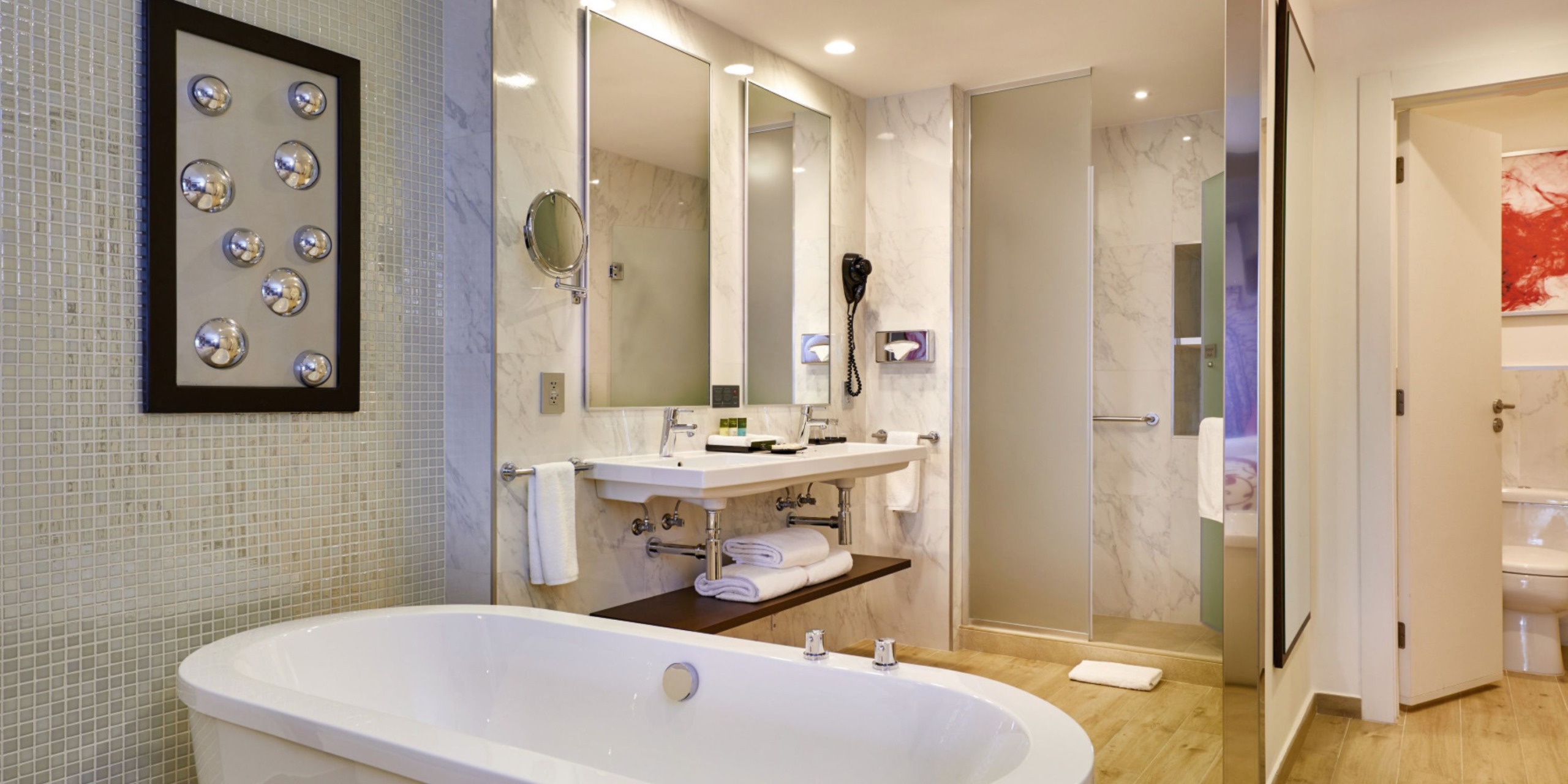 Experience the modern bathroom at Hotel Riu Palace Costa Mujeres, featuring a freestanding bathtub, double sinks, wall-mounted hairdryer, and a glass-enclosed shower. The marble walls and floor perfectly complement the contemporary decor.