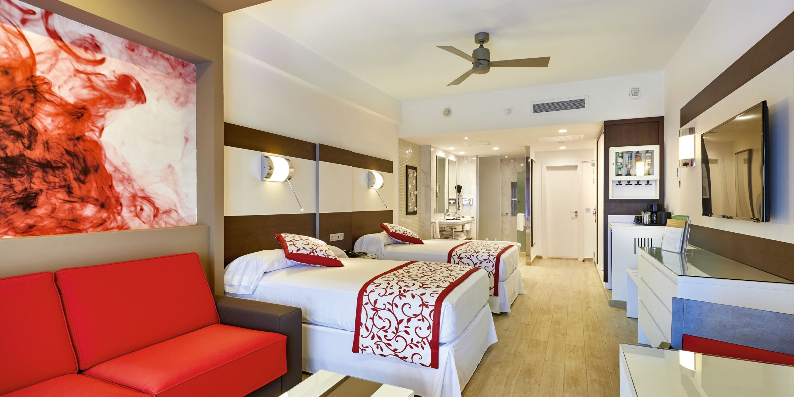 The Hotel Riu Palace Costa Mujeres offers modern rooms featuring two beds, red and white decor, a ceiling fan, a wall-mounted TV, and a seating area with a red sofa.