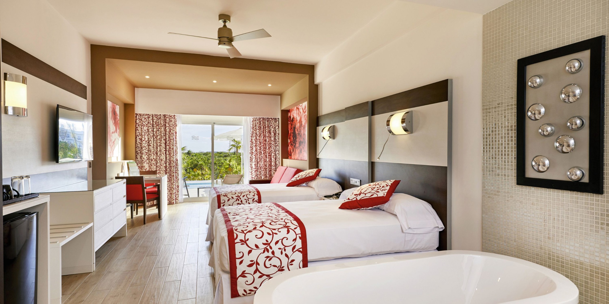 A room at Hotel Riu Palace Costa Mujeres features two twin beds, a decorative chair, wall-mounted lamps, a flat-screen TV, a ceiling fan, and a freestanding bathtub. It also offers a balcony view overlooking lush greenery.