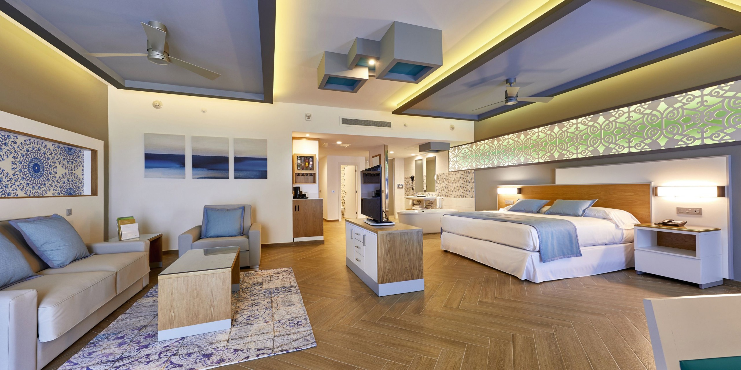 Enjoy a spacious room at Hotel Riu Palace Costa Mujeres, featuring a large bed, seating area, flat-screen TV, and modern decor. The room is enhanced with soft lighting and a stylish patterned wall accent.