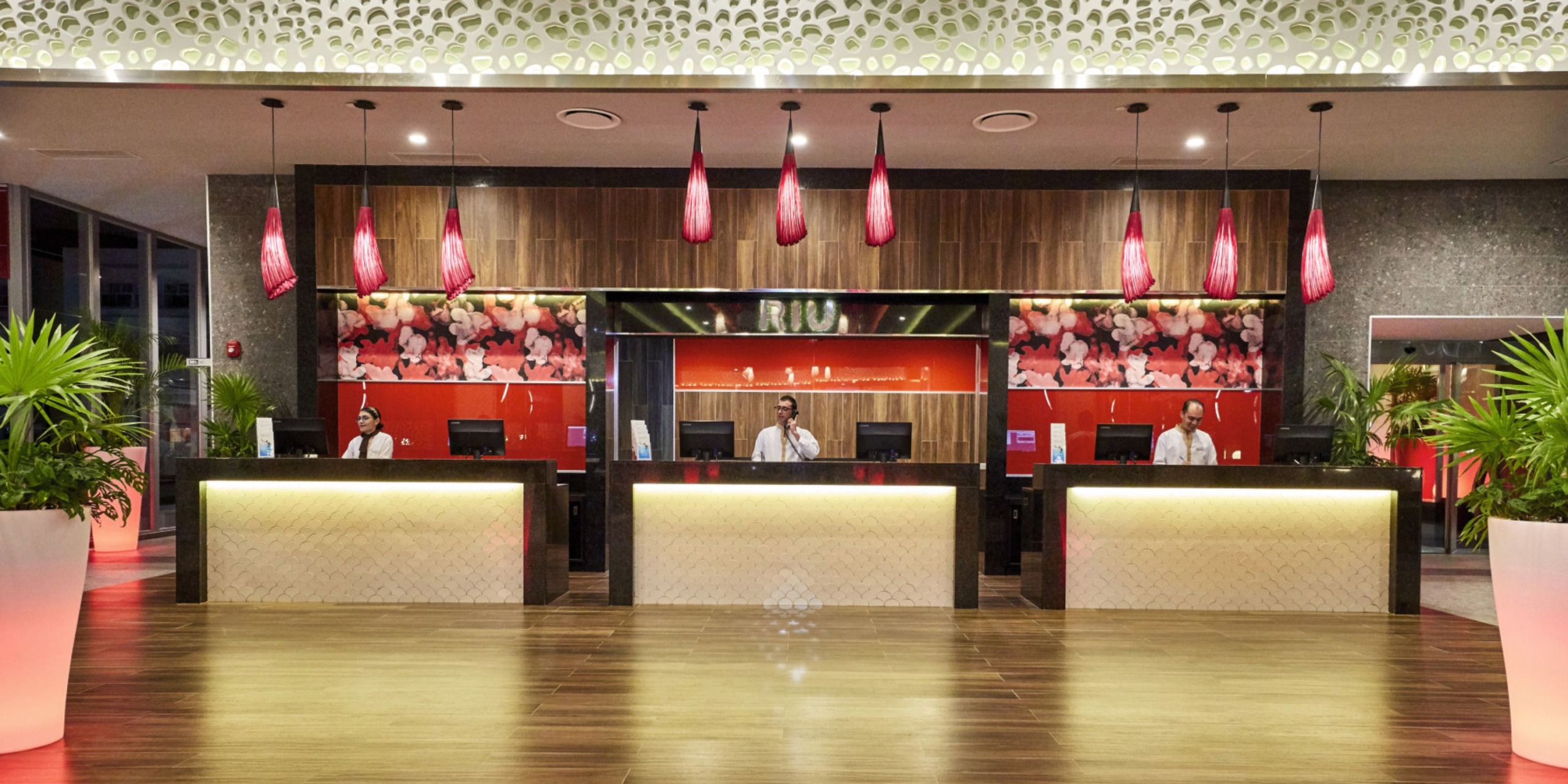 The reception area of Hotel Riu Palace Costa Mujeres features three counters, eye-catching red hanging lights, and tropical plant decor, with three attentive staff members standing behind the counters.