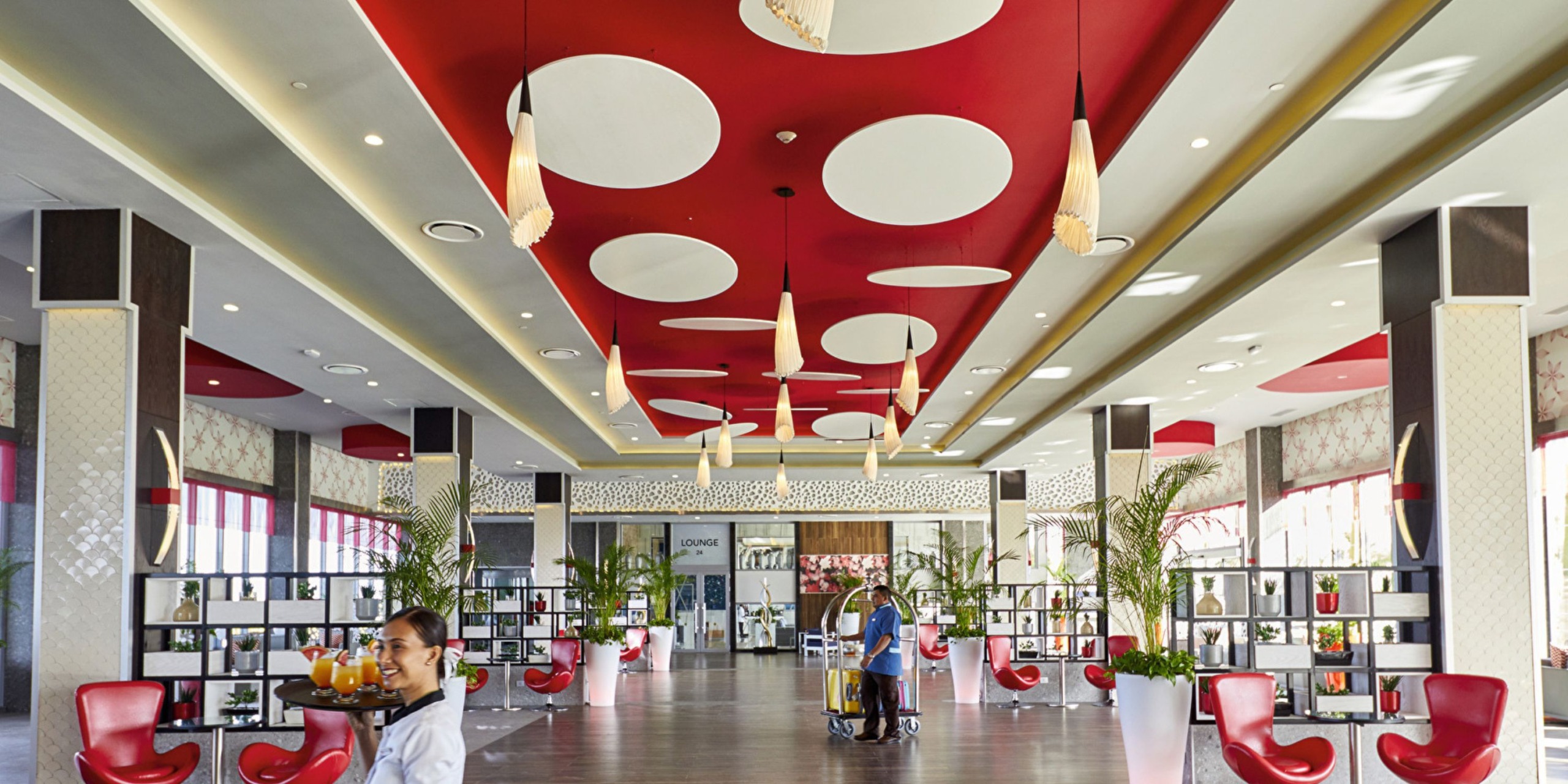 The contemporary lobby of Hotel Riu Palace Costa Mujeres features a striking red and white ceiling design, pendant lighting, large potted plants, and a cozy seating area. One guest carries a tray of drinks while another navigates the space with their luggage in tow.