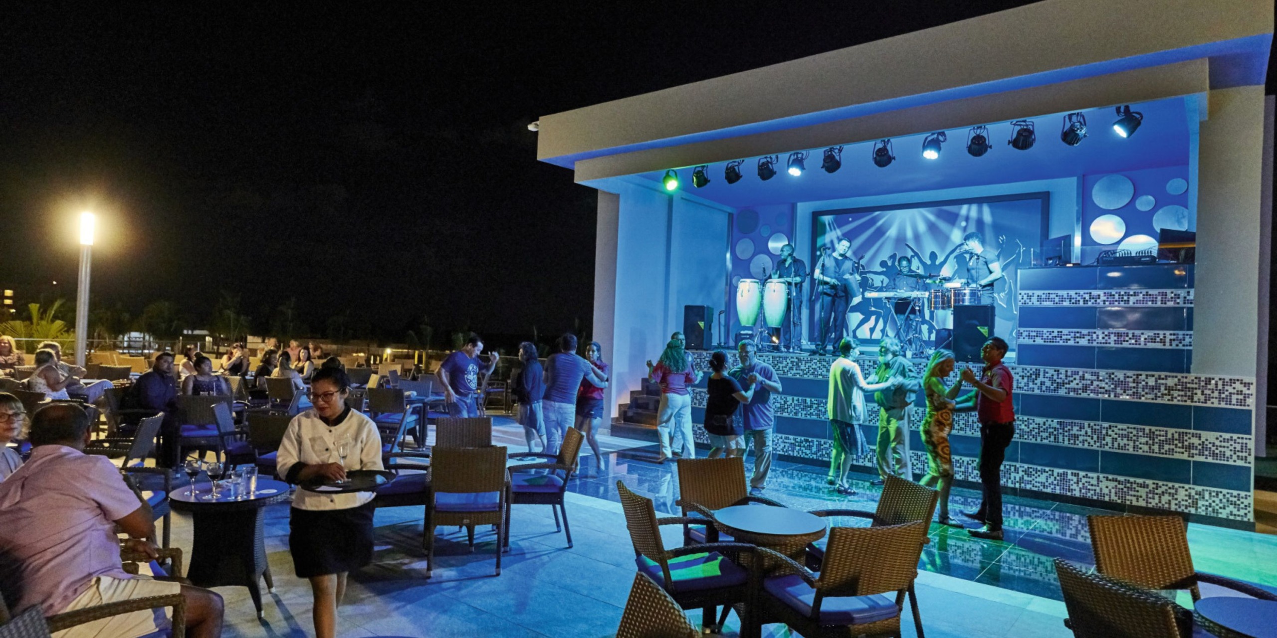 Experience live outdoor music performances at Hotel Riu Palace Costa Mujeres, where guests dine and dance under the night sky. Bright stage lights illuminate the performers as tables and chairs are thoughtfully arranged in the foreground.