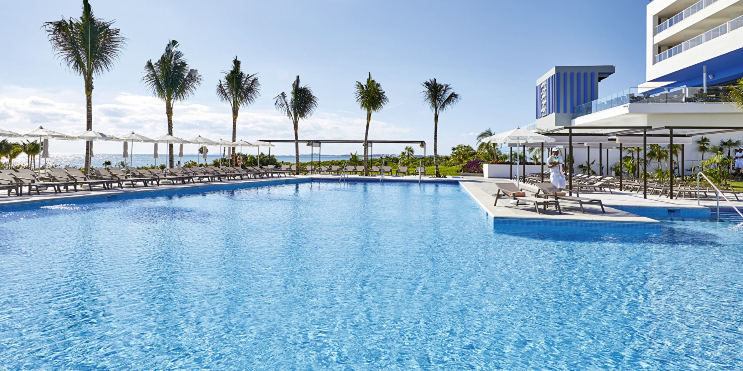 Experience the outdoor pool area at Hotel Riu Palace Costa Mujeres, featuring lounge chairs and umbrellas surrounded by palm trees and a modern building, with the ocean visible in the background under a clear blue sky.