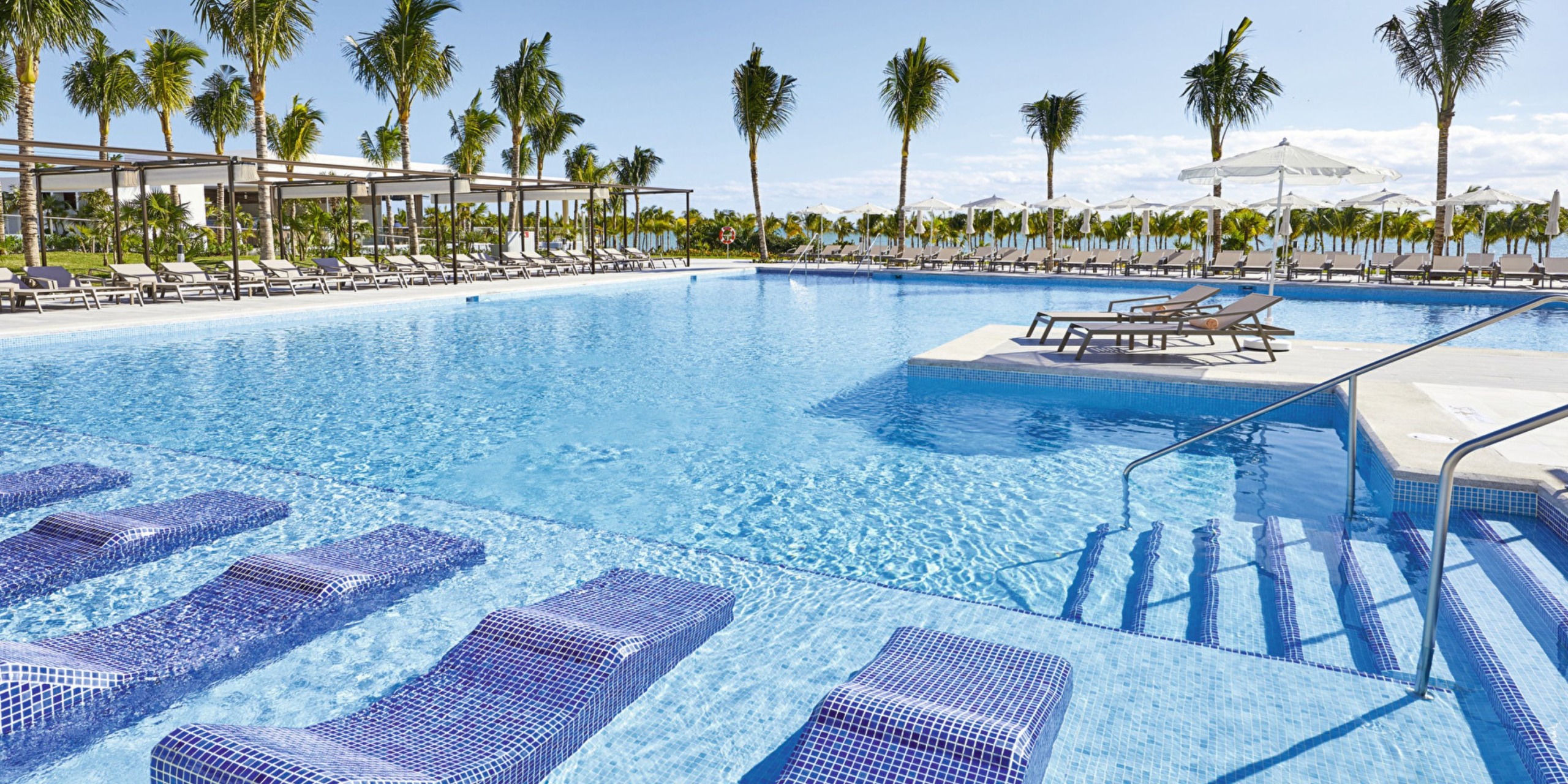 The Hotel Riu Palace Costa Mujeres features an outdoor swimming pool with tiled submerged loungers, encircled by palm trees and deck chairs set under a clear blue sky.