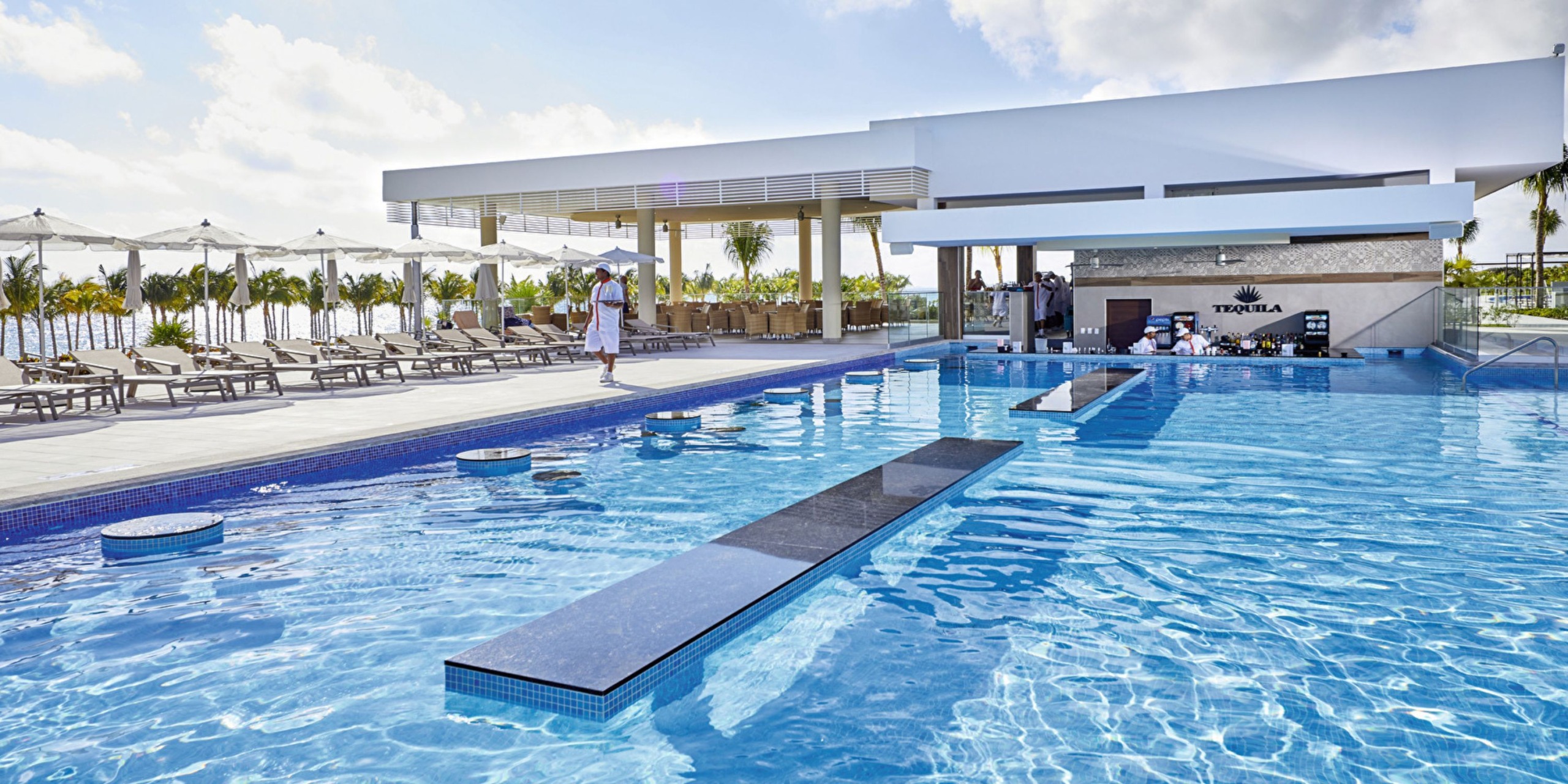 Pool area at Hotel Riu Palace Costa Mujeres featuring a swim-up bar, lounging chairs, umbrellas, and a clear sky.
