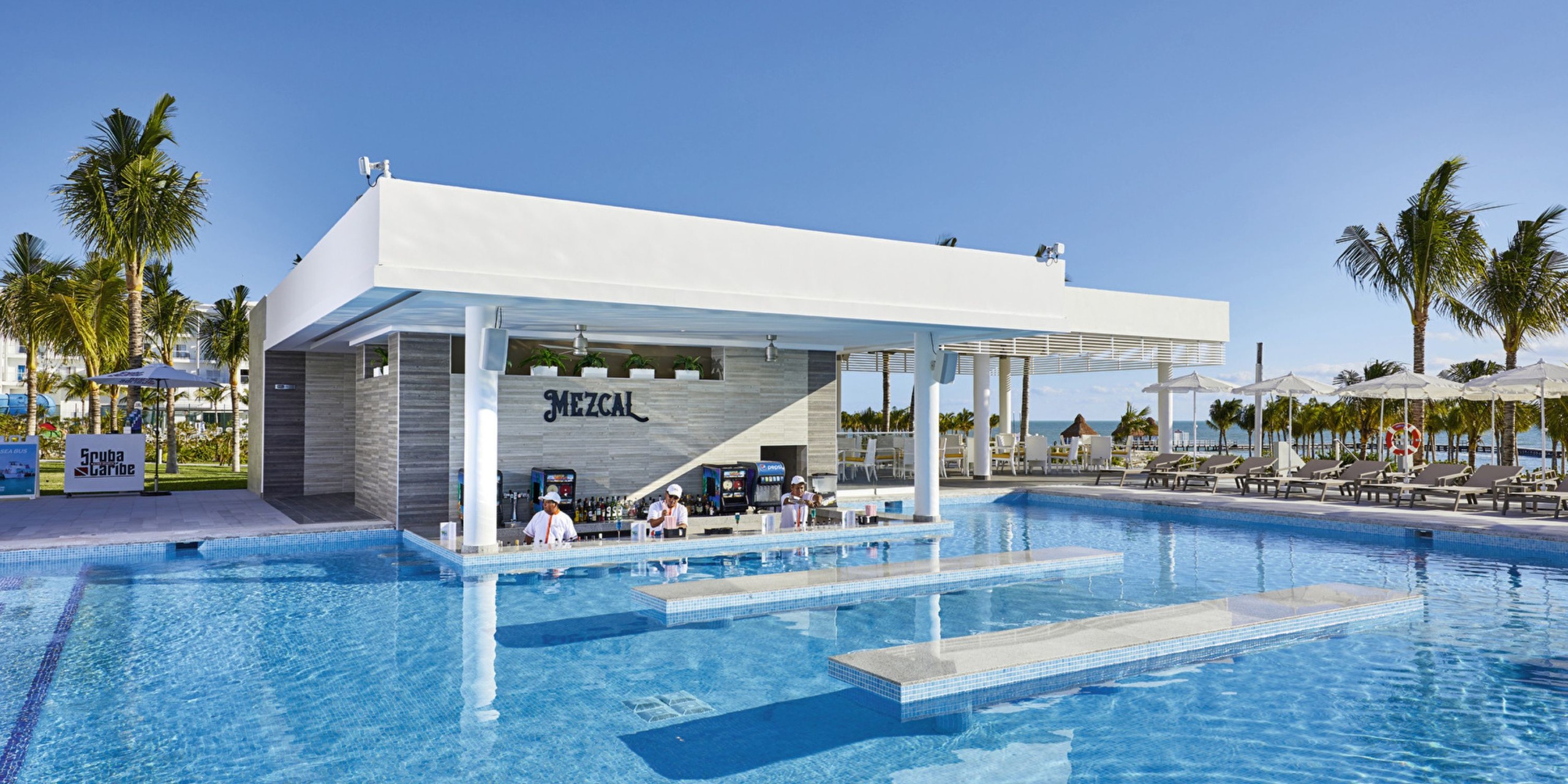The Hotel Riu Palace Costa Mujeres features an outdoor swim-up bar with stools in a spacious pool. Palm trees and lounge chairs are visible around the area, set against a clear and sunny sky.