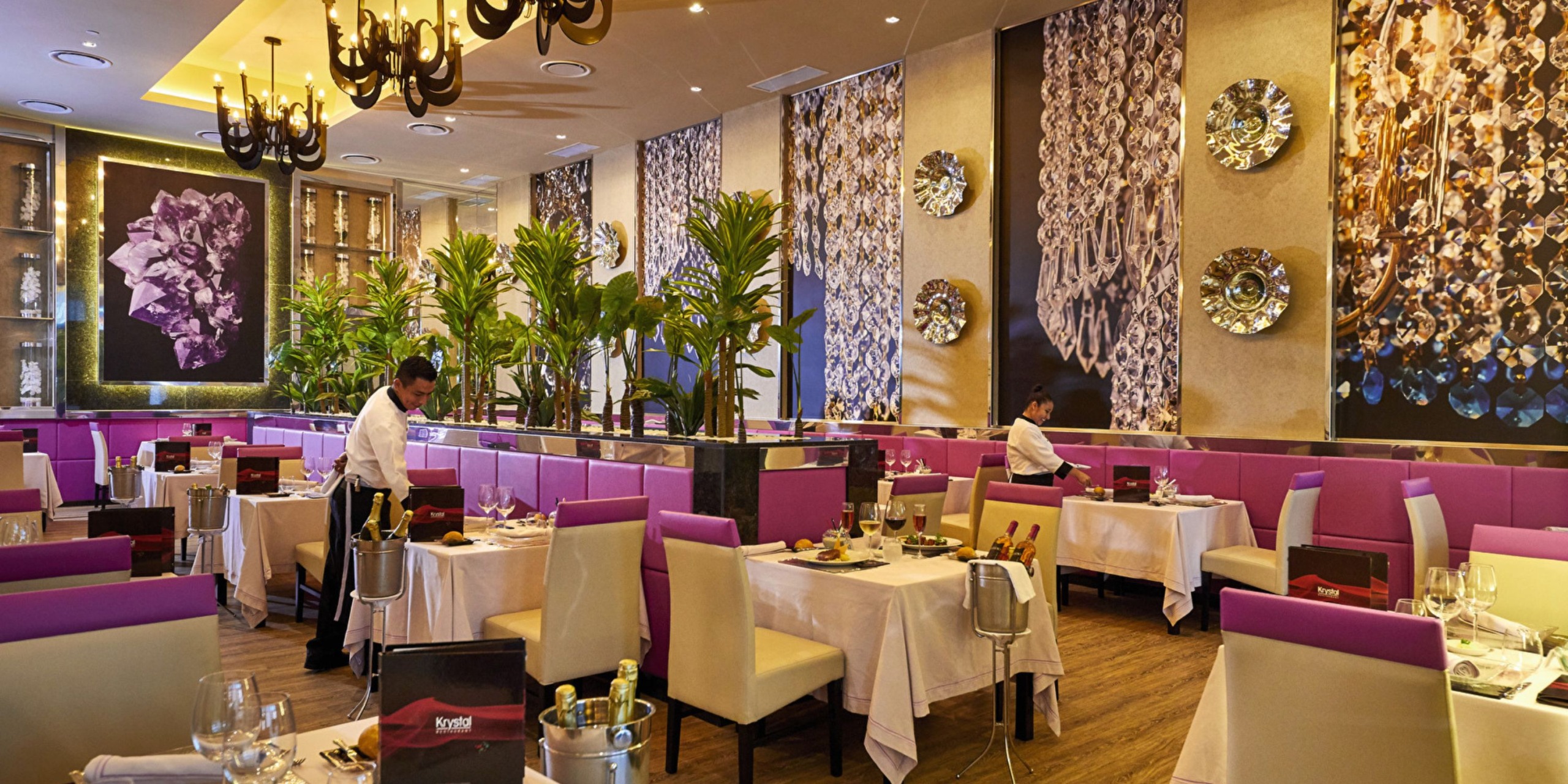 The sophisticated interior of Hotel Riu Palace Costa Mujeres features elegant pink booths, pristine white tablecloths, opulent chandeliers, and captivating wall art, as two staff members attentively set the tables.