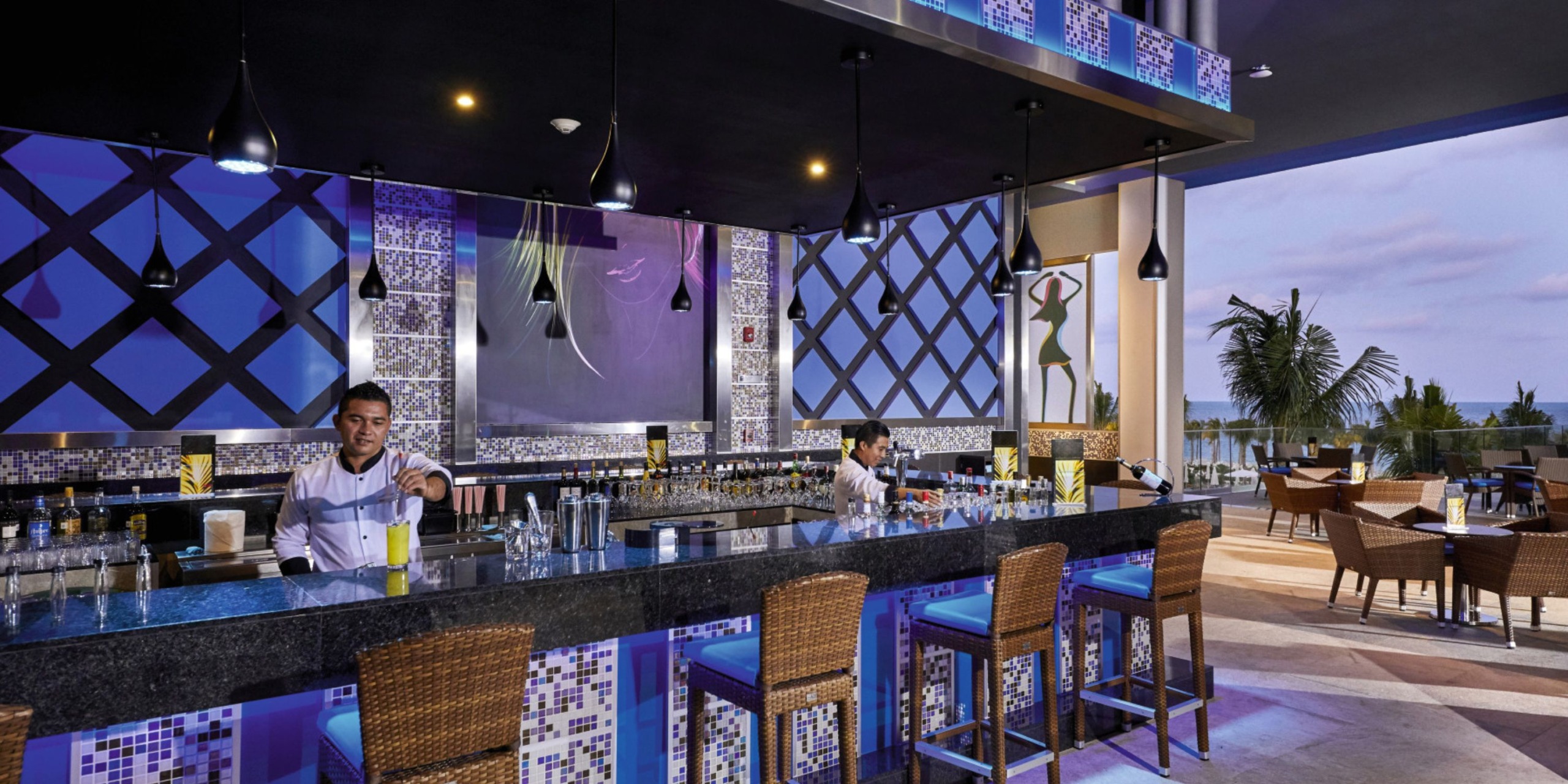 Bartenders working behind a chic outdoor bar at Hotel Riu Palace Costa Mujeres, featuring wicker stools, mosaic tile accents, and overlooking palm trees and the ocean.