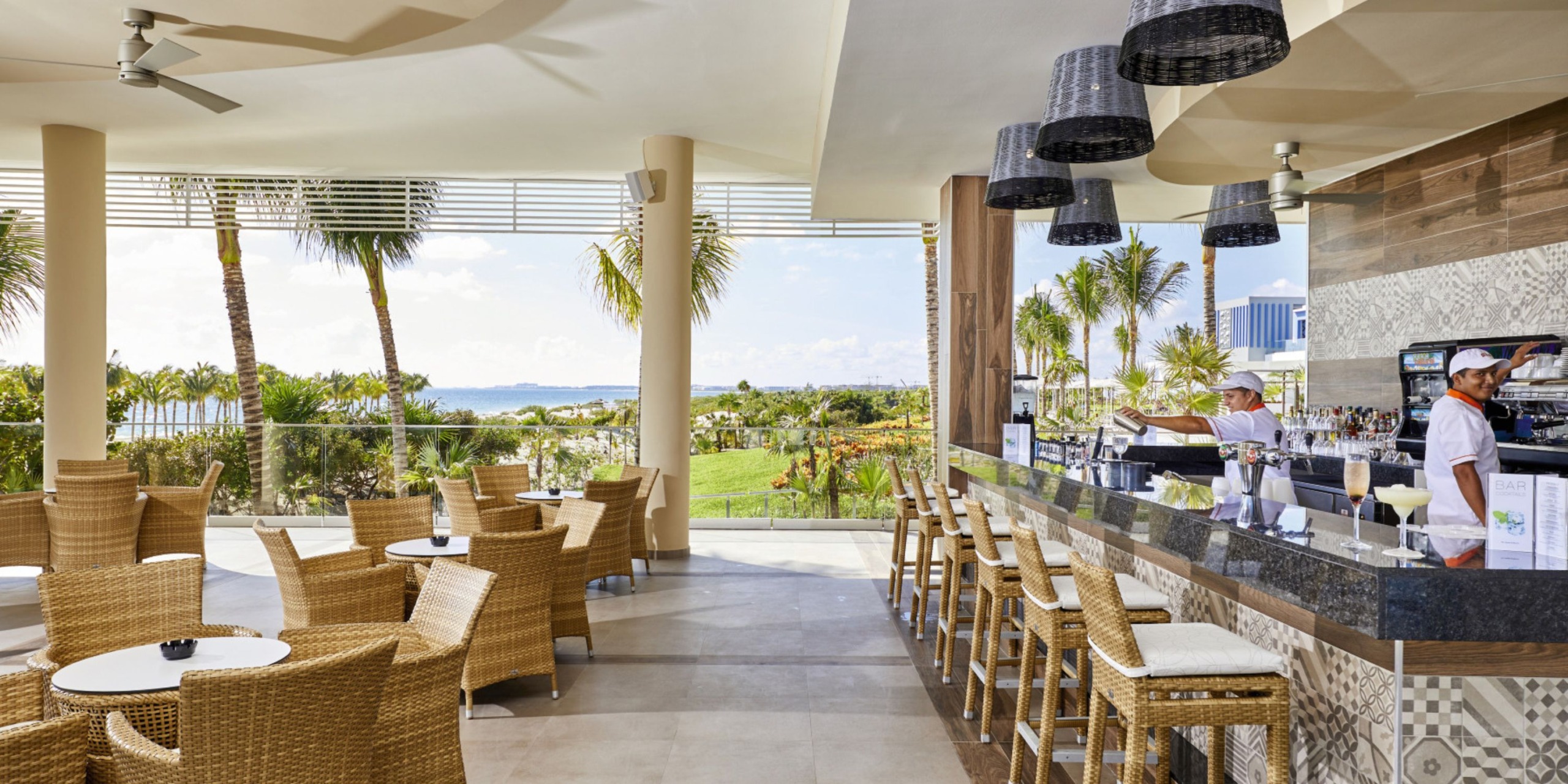 The Hotel Riu Palace Costa Mujeres features an outdoor café with wicker chairs and tables, a bar counter with stools, and palm trees in the background. Two staff members are stationed behind the bar.
