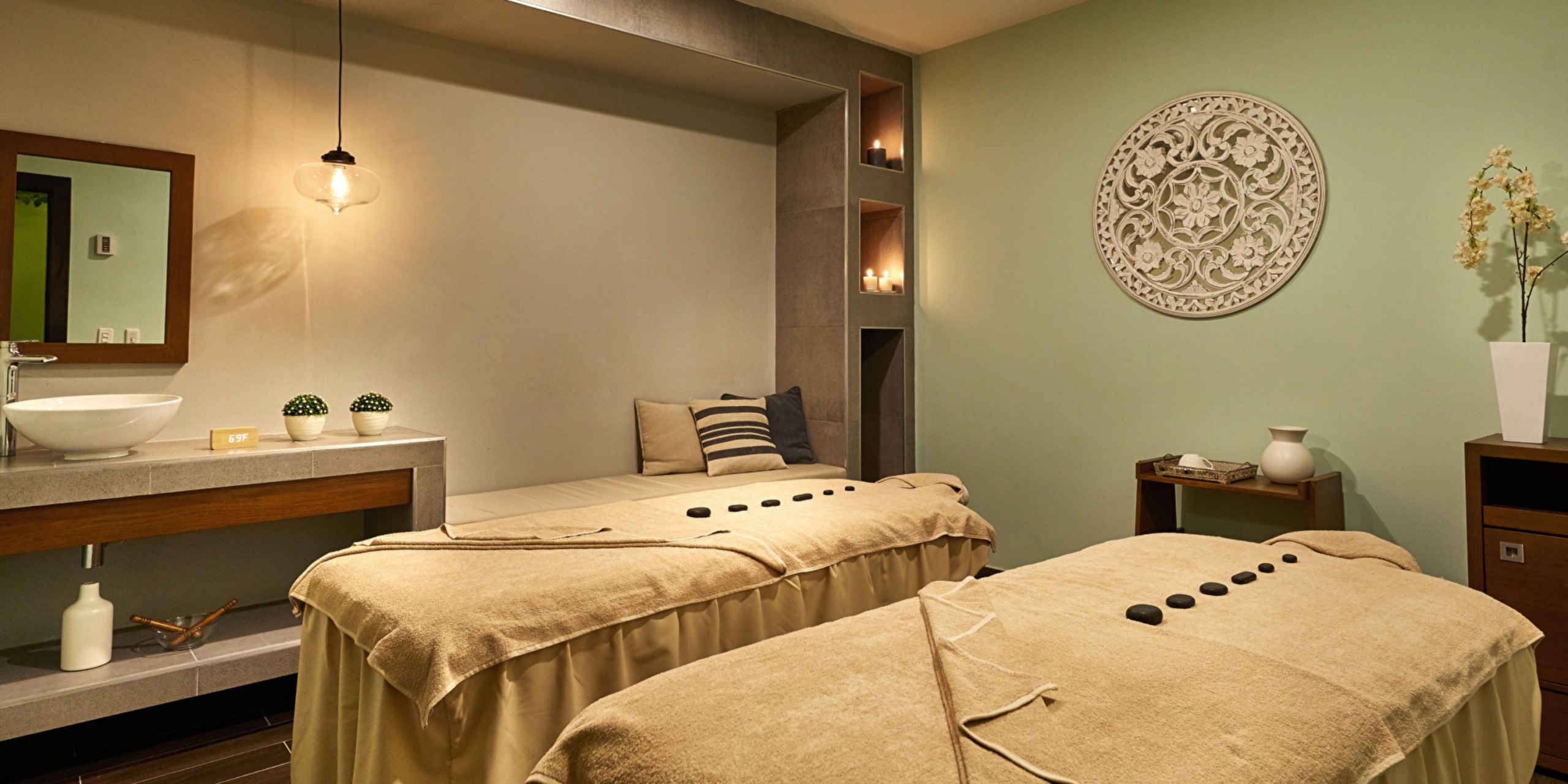 A tranquil spa room at the Hotel Riu Palace Costa Mujeres features two massage tables, each adorned with towels and hot stones, while soft lighting and a decorative wall piece enhance the relaxing ambiance.