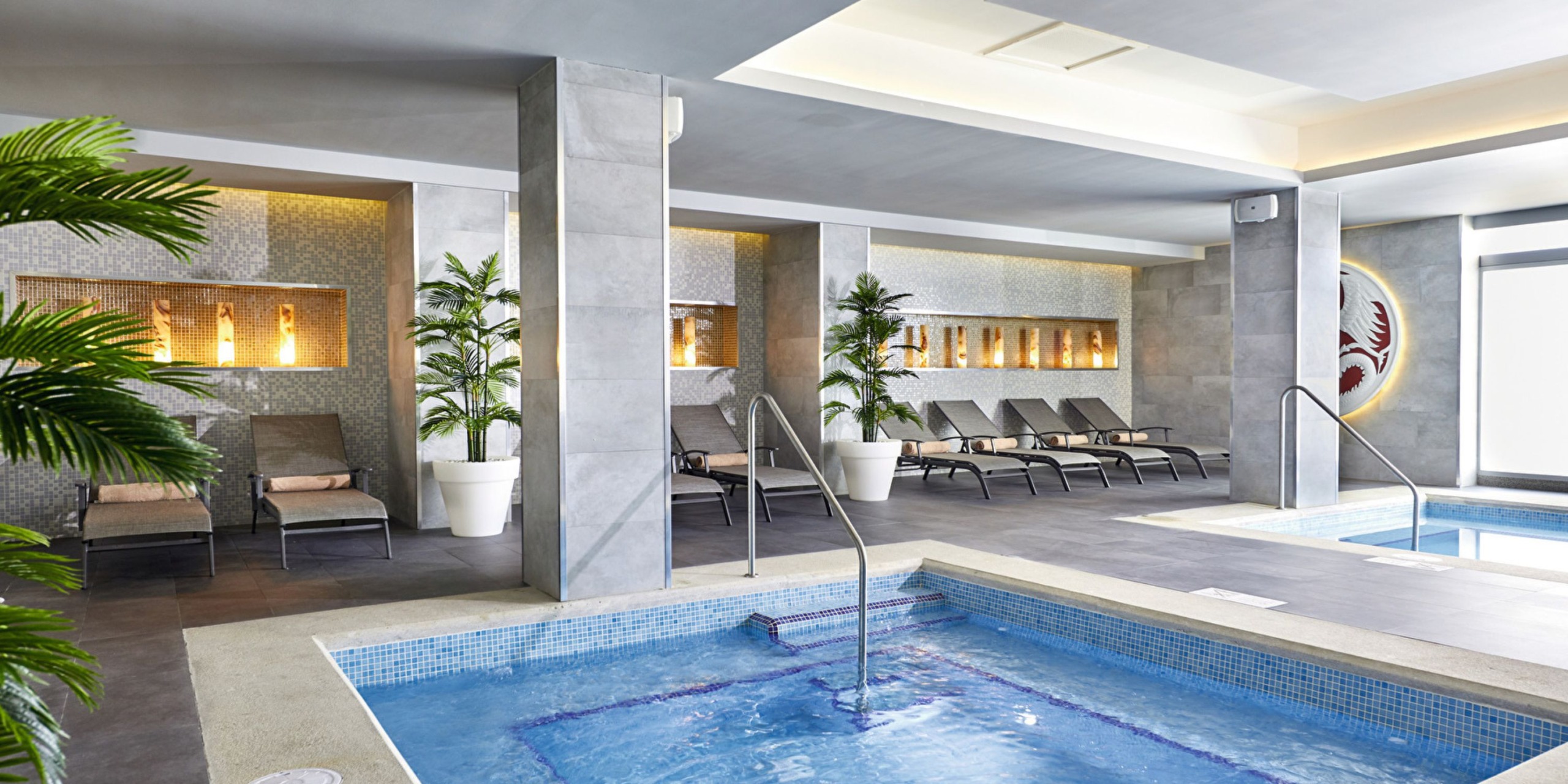 The indoor spa area at Hotel Riu Palace Costa Mujeres includes a small pool, lounge chairs, potted plants, and features a modern design with neutral tones and recessed lighting.