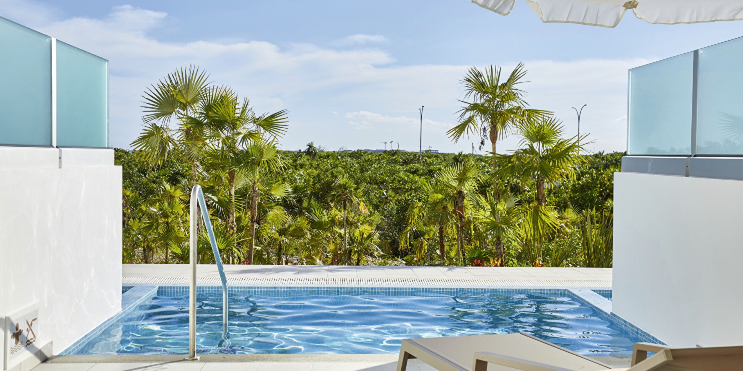 A small rectangular pool with clear water at Hotel Riu Palace Costa Mujeres is surrounded by white walls and palm trees in the background, with two sun loungers positioned in the foreground under a large white umbrella.