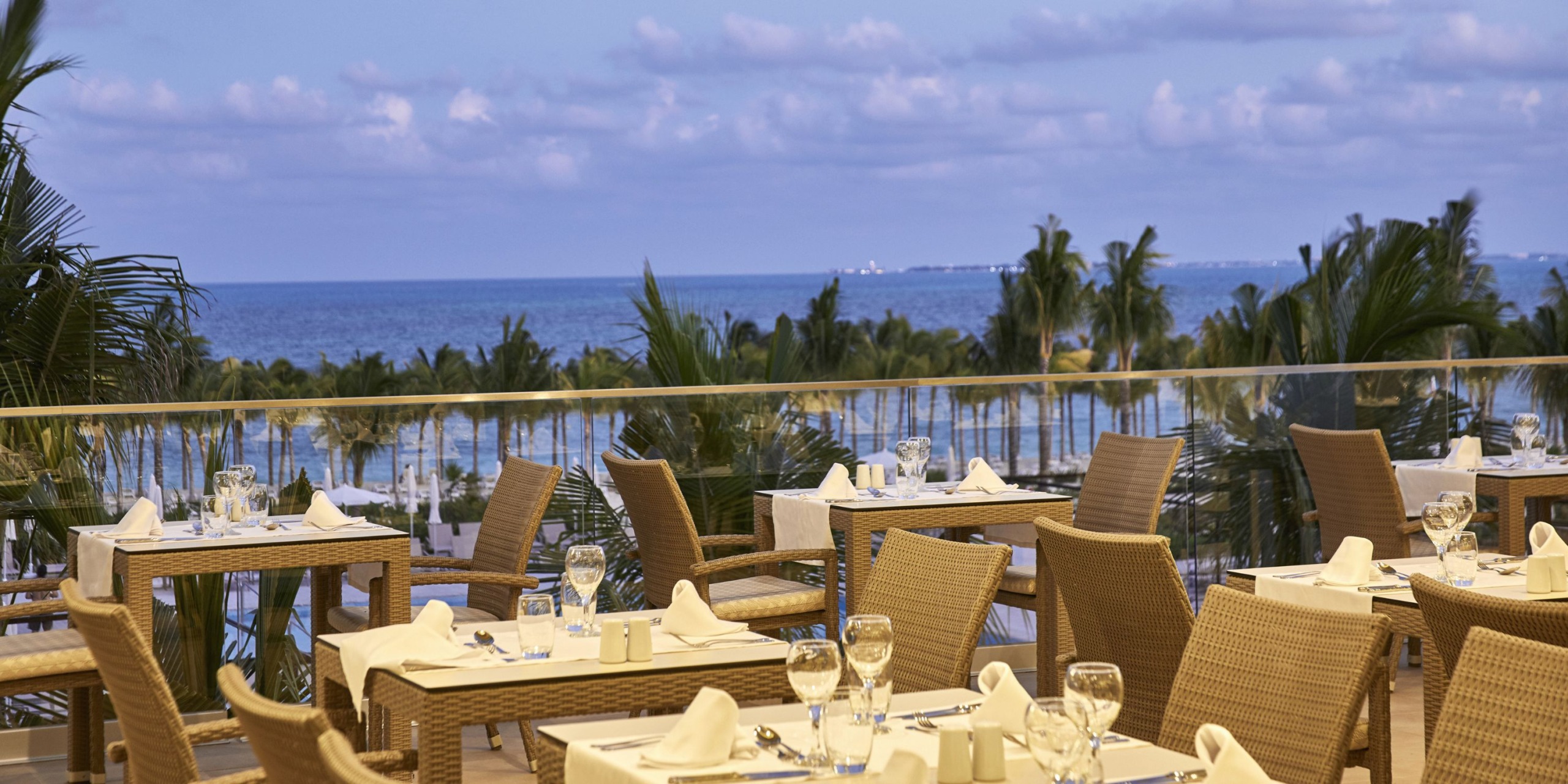 Outdoor restaurant at Hotel Riu Palace Costa Mujeres featuring wicker chairs and tables elegantly set with white tablecloths, napkins, and glassware. Palm trees adorn the landscape, complemented by a breathtaking ocean view in the background.
