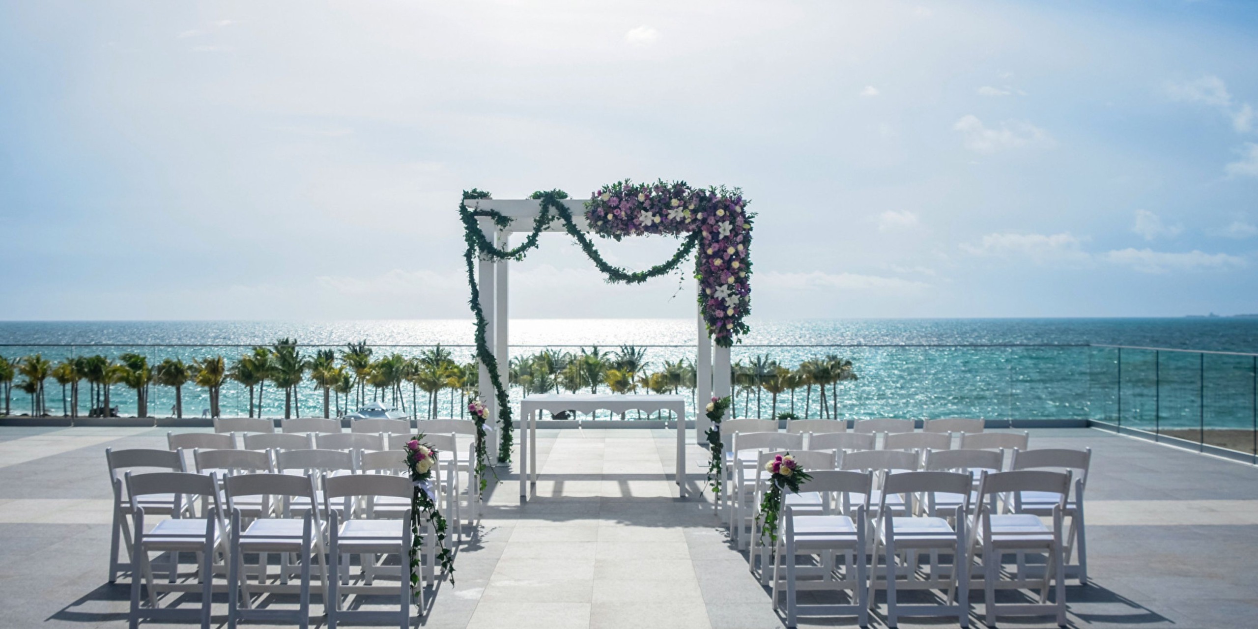 An outdoor wedding setup overlooking the sea at Hotel Riu Palace Costa Mujeres, featuring white chairs and a floral arch under a clear sky.