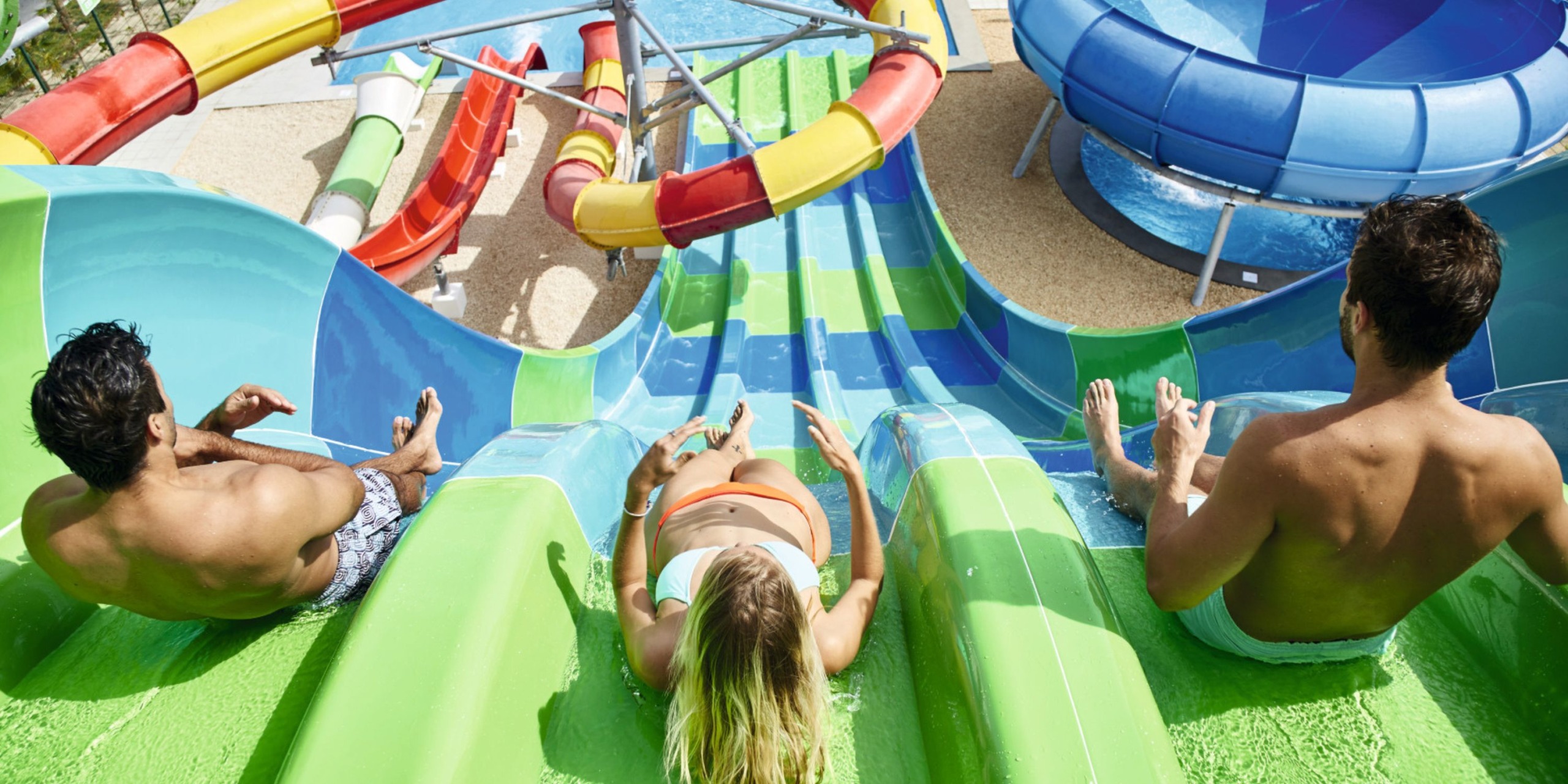 Three guests at the Hotel Riu Palace Costa Mujeres are enjoying the vibrant water slides, with one person in the center who has green hair and two others gliding alongside.