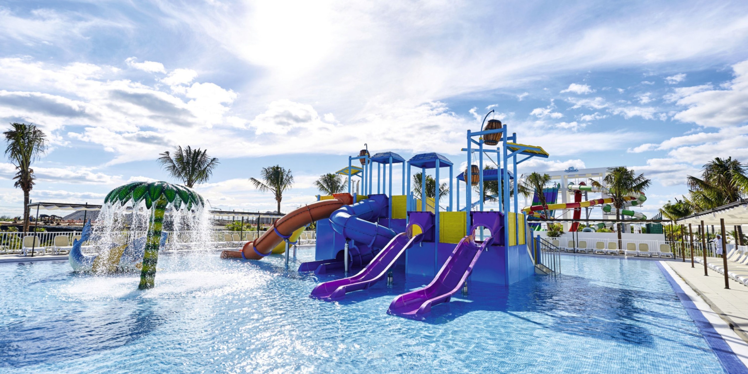 The Hotel Riu Palace Costa Mujeres features a vibrant water playground complete with slides and splash elements set in a pool beneath a sunny sky, with palm trees serving as a scenic backdrop.