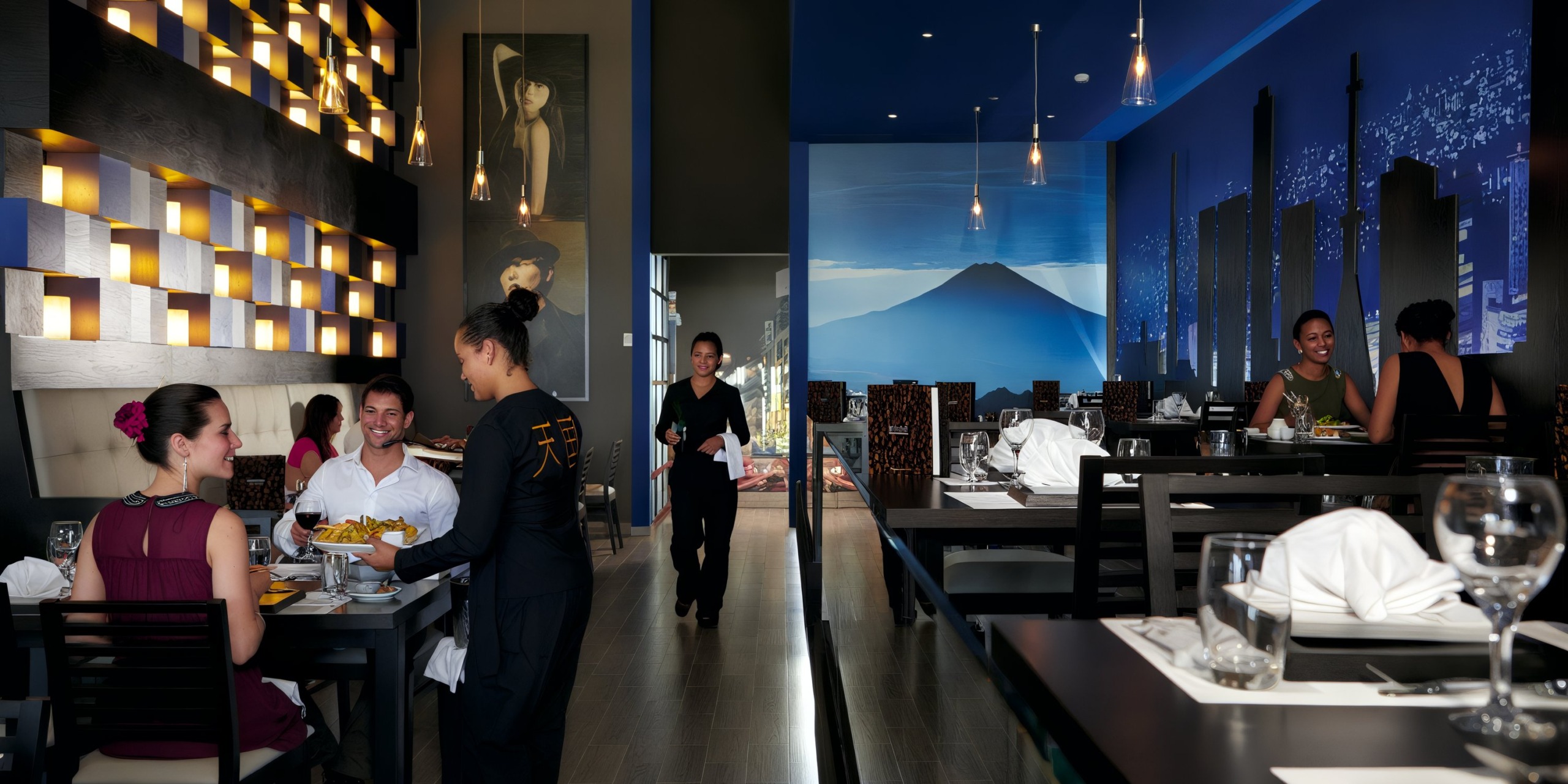 A contemporary interior at Hotel Riu Palace Costa Rica features dark wood furniture, with one server engaging guests and another walking by. The blue-lit wall is adorned with a large mural depicting a mountain and skyline.