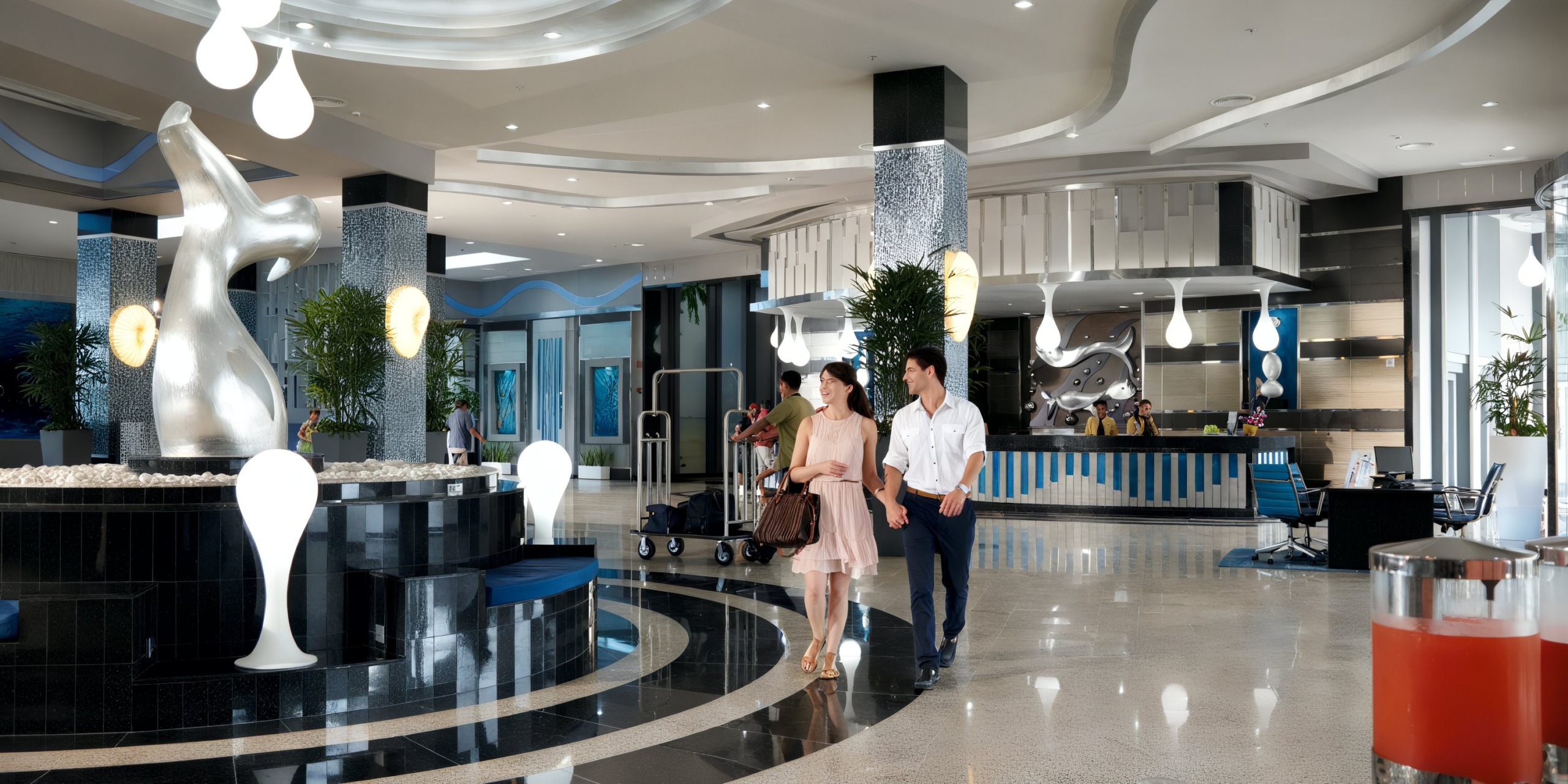 A couple strolls through the contemporary lobby of Hotel Riu Palace Costa Rica, featuring abstract sculptures, sweeping design elements, and a reception desk in the background.