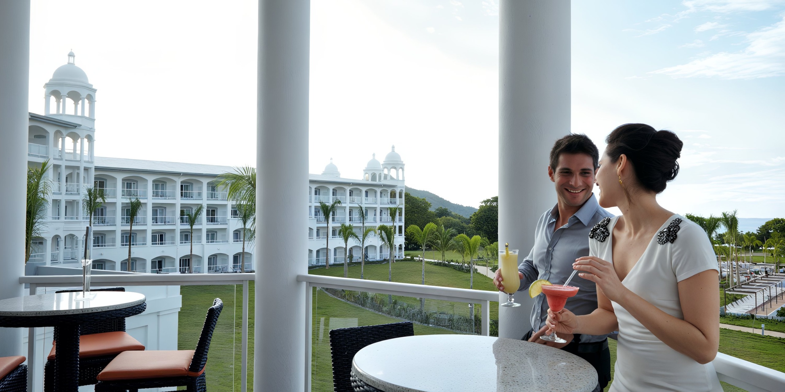 A man and woman enjoy drinks on a balcony overlooking the Hotel Riu Palace Costa Rica, featuring its distinctive towers and lush garden.