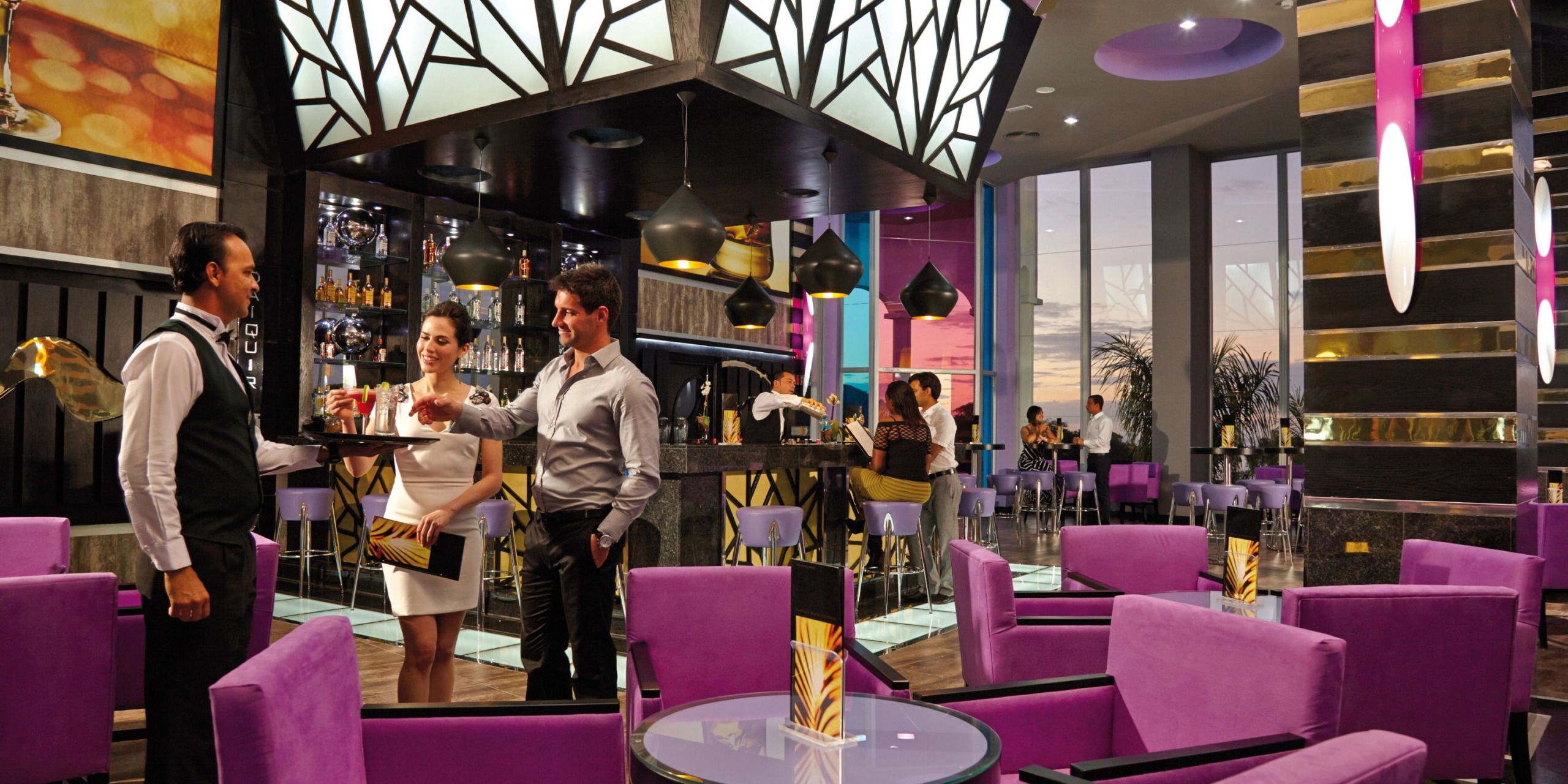 Guests are socializing at the Hotel Riu Palace Costa Rica, where purple seating, geometric lighting, and a well-stocked bar area create a modern atmosphere. The sunset can be seen through large windows.