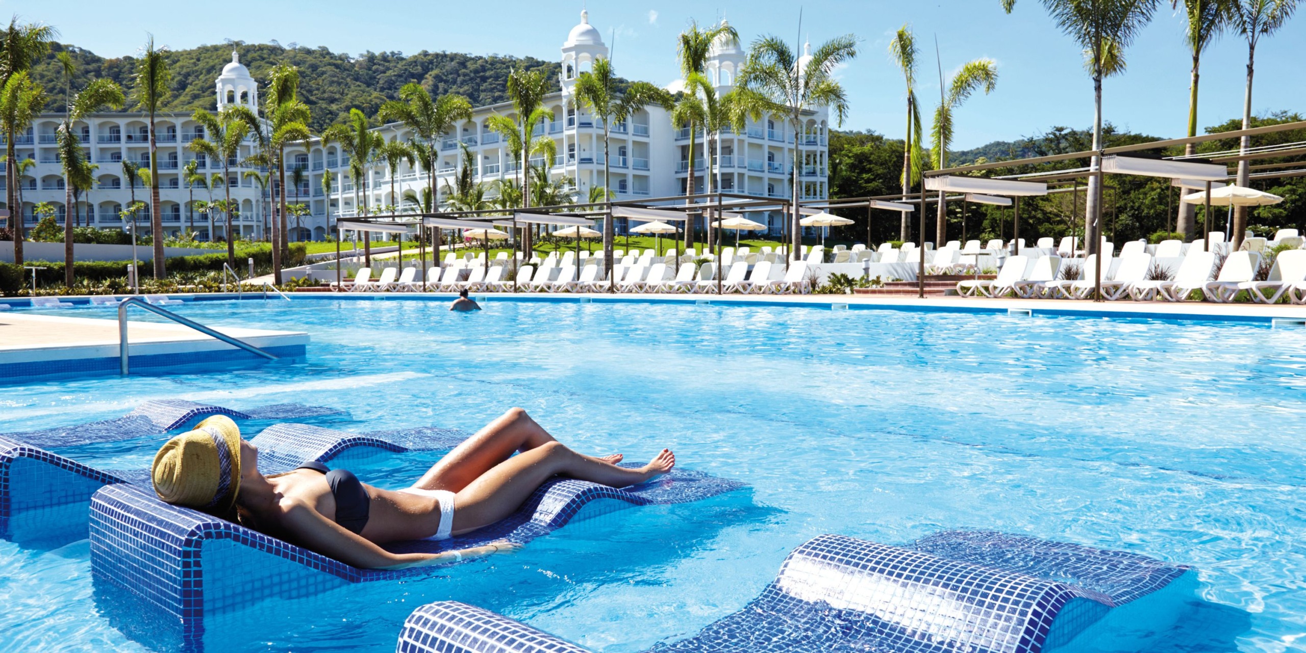 A person relaxes on a lounger in the expansive outdoor pool at Hotel Riu Palace Costa Rica, surrounded by palm trees with the majestic white hotel building as a backdrop.