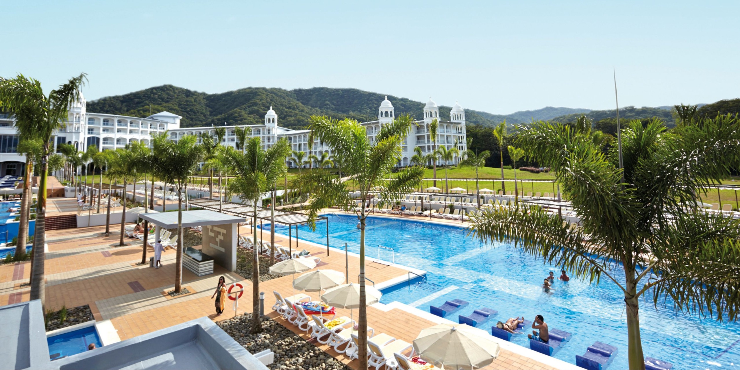 Hotel Riu Palace Costa Rica features a spacious swimming pool surrounded by sun loungers and palm trees. The background showcases white buildings with a stunning vista of distant hills beneath a clear sky.