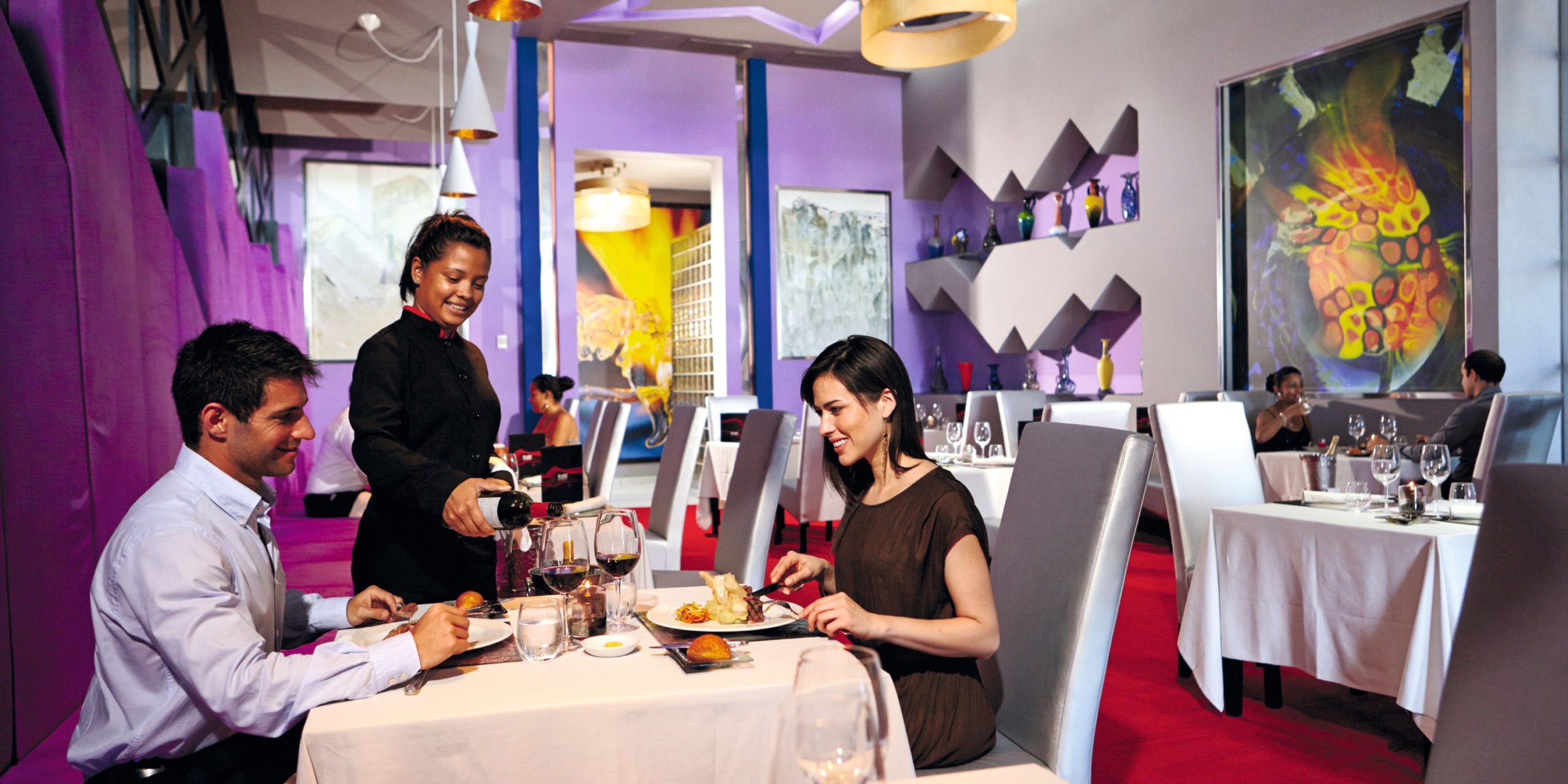 At Hotel Riu Palace Costa Rica, a server pours wine for a couple seated at a table in a modern restaurant adorned with purple walls and contemporary art.