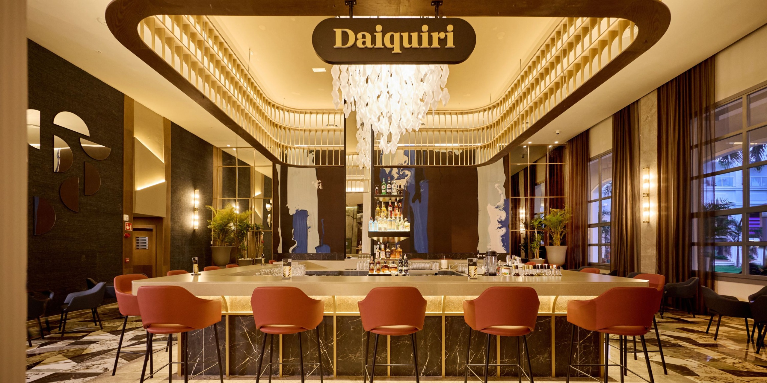 A contemporary bar named "Daiquiri" at the Hotel Riu Palace Bavaro, featuring a chandelier, surrounded by red chairs and a marble countertop.