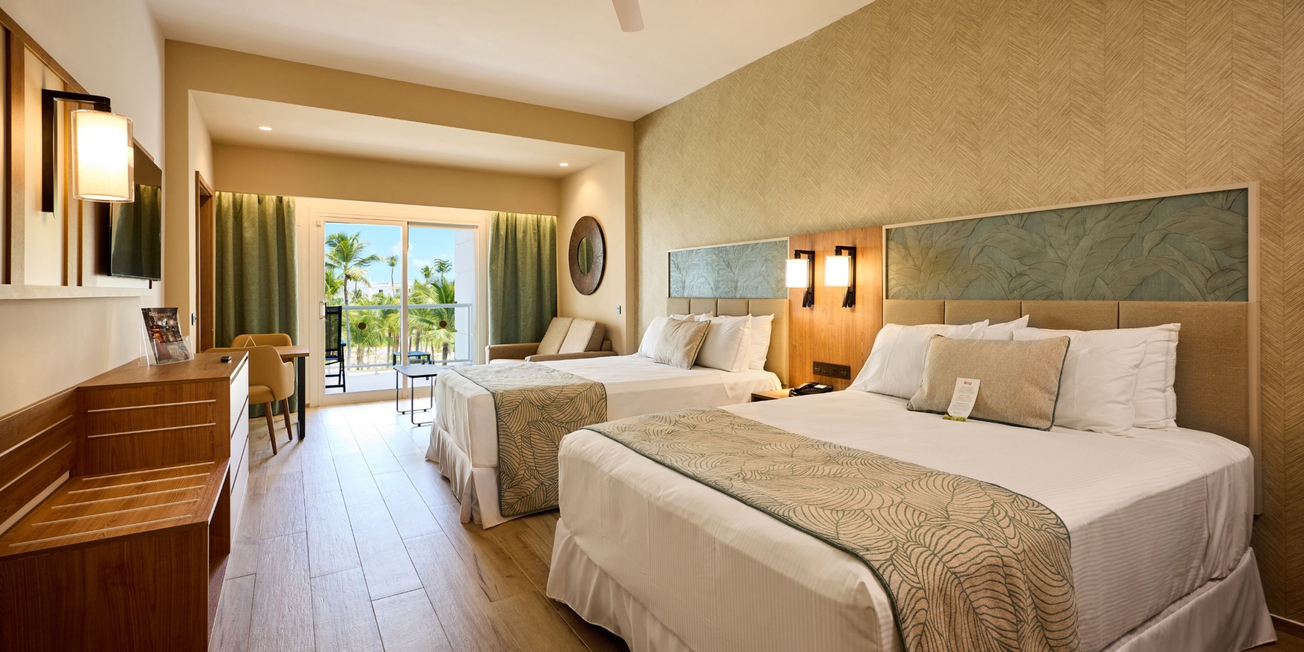 The Hotel Riu Palace Bavaro offers a room featuring two double beds adorned with decorative pillows, a desk, and a sliding door that opens to a balcony overlooking palm trees. The room showcases a neutral color palette and wooden flooring.