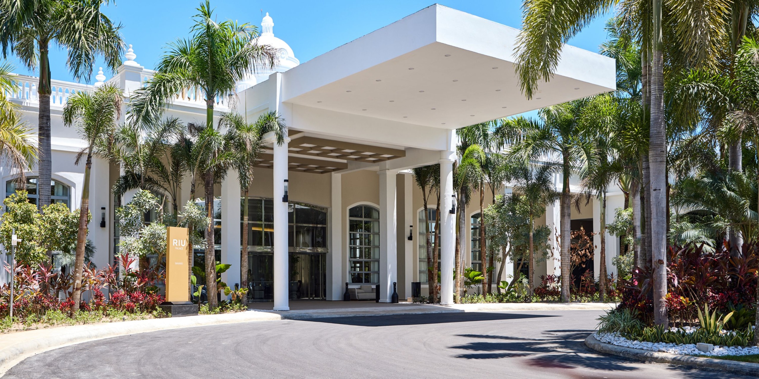The entrance of Hotel Riu Palace Bavaro features a grand facade with tall columns and a flat roof, surrounded by palm trees and vibrant plants.