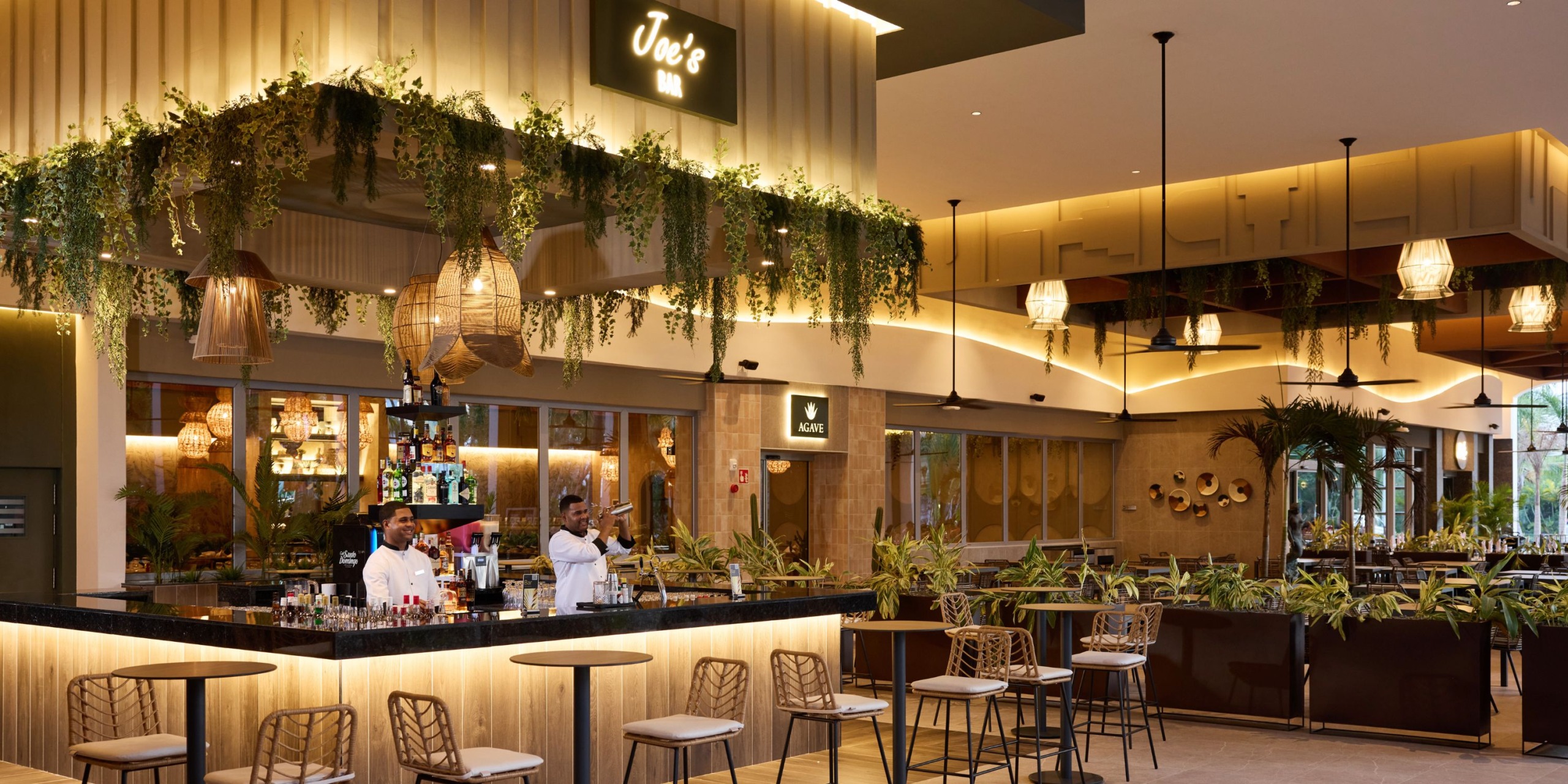 The interior of Hotel Riu Palace Bavaro features a contemporary bar with two staff members behind the counter, elegantly surrounded by decorative plants, ample seating, and stylish pendant lights.