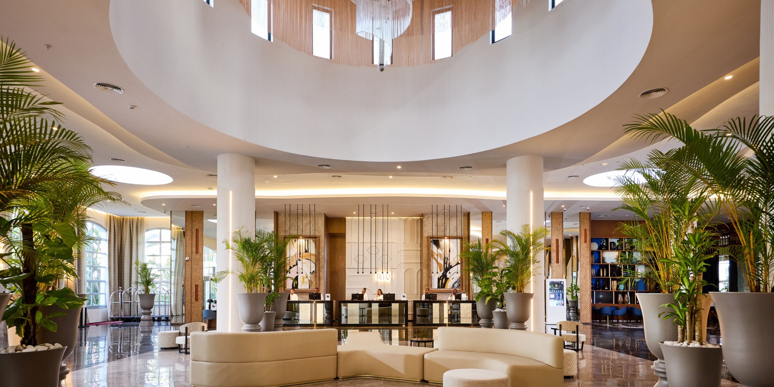 The spacious lobby of the Hotel Riu Palace Bavaro features large circular seating, tall white columns, potted plants, and a reception desk set against a backdrop of elevators, with natural light streaming in from upper windows.