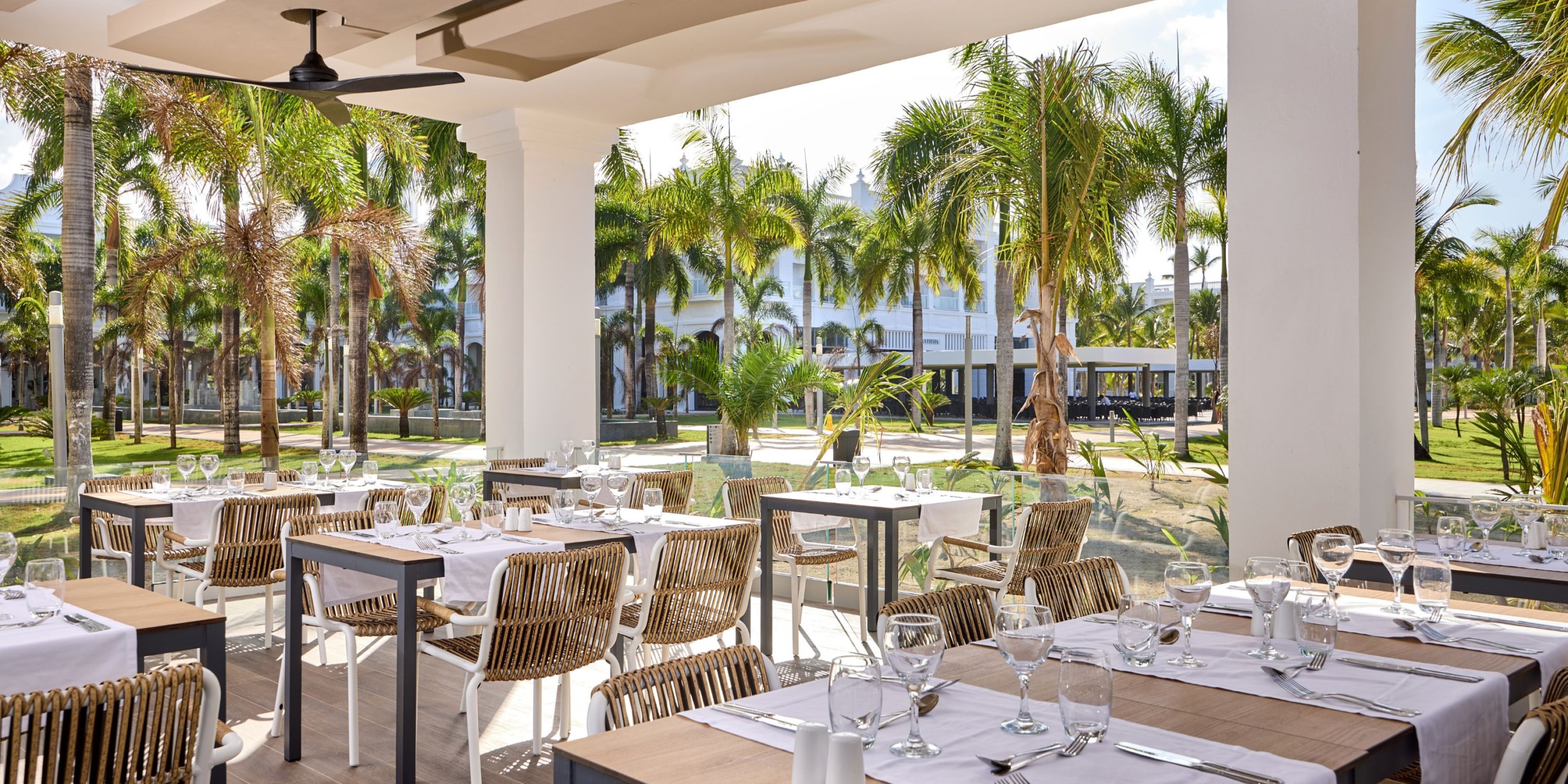Outdoor dining area at Hotel Riu Palace Bavaro, featuring neatly arranged tables and chairs, with a view of a garden adorned with palm trees.