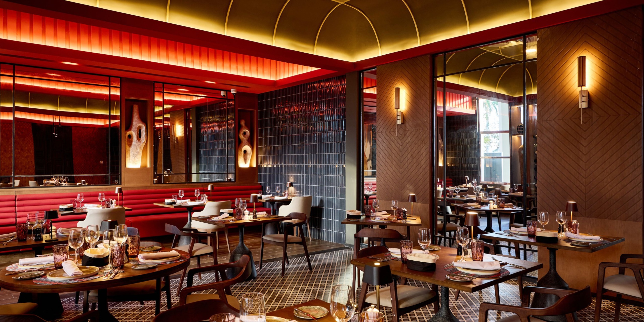 The Hotel Riu Palace Bavaro boasts a stylish interior with modern decor, featuring red booths, wooden tables, and elegant lighting. Each table is beautifully set with plates and glasses.