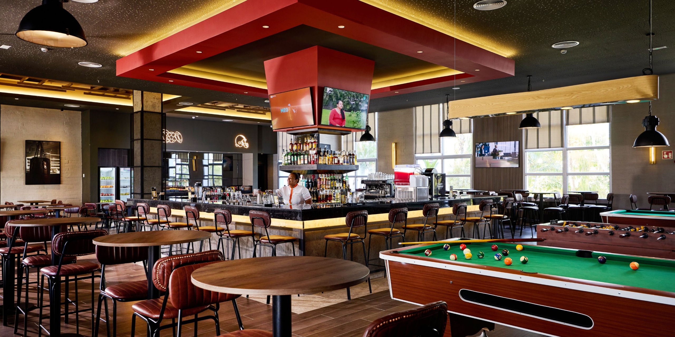 Hotel Riu Palace Bavaro features a lively bar with round tables, stools, a central counter, and a pool table. A staff member stands behind the counter ready to serve guests. Overhead TVs provide entertainment as natural light streams through the large windows.