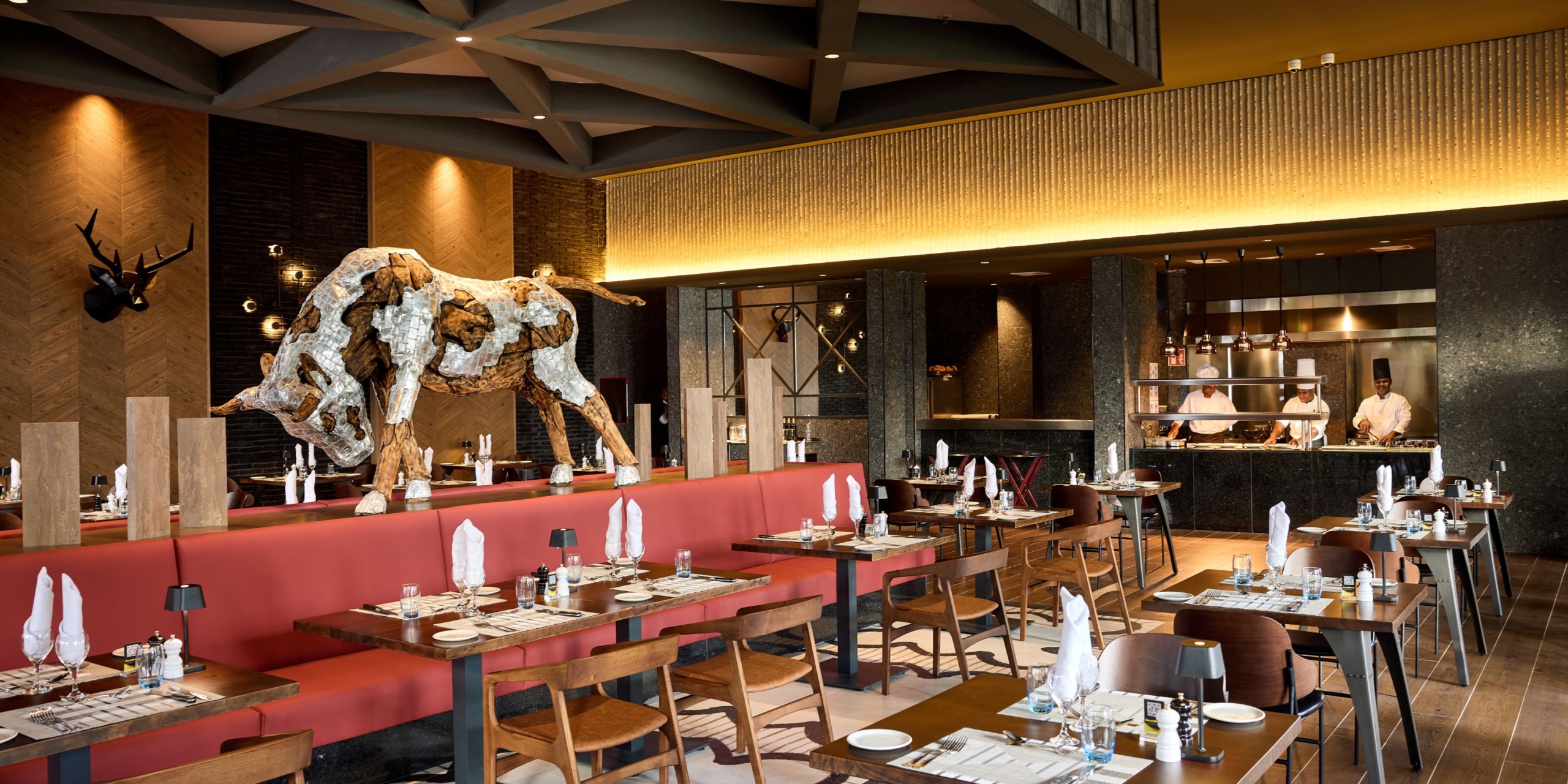 Stylish interior at Hotel Riu Palace Bavaro featuring wooden tables, red booth seating, and a prominent decorative bull sculpture. The open kitchen allows guests to watch chefs in action.