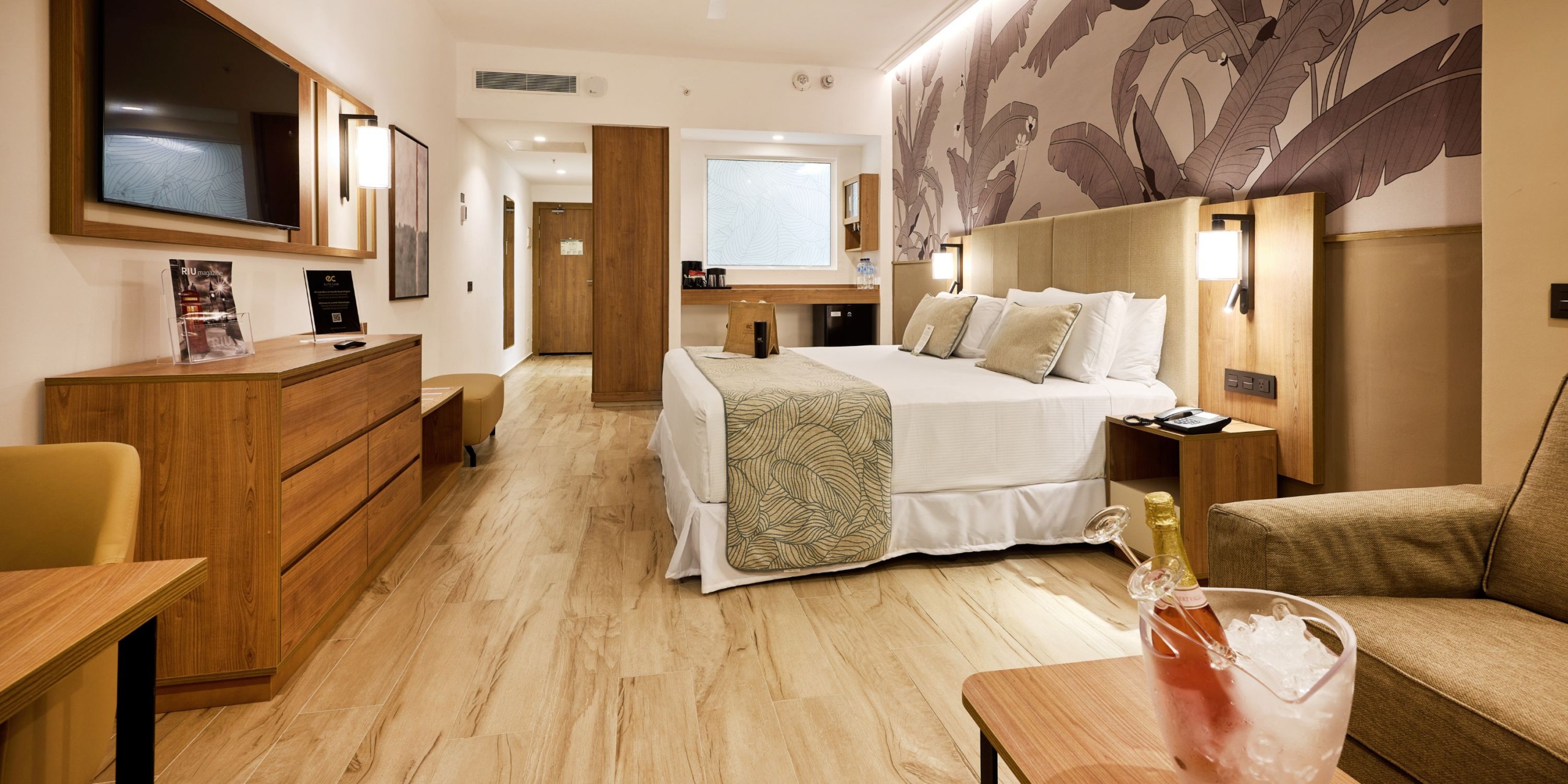 Experience the contemporary comfort of a Hotel Riu Palace Bavaro room, featuring a spacious bed and elegant wood furnishings, accented by a stylish decorative wall. In the foreground, find a table with a refreshing drink chilling in an ice bucket, while a mirror enhances the space from the background.