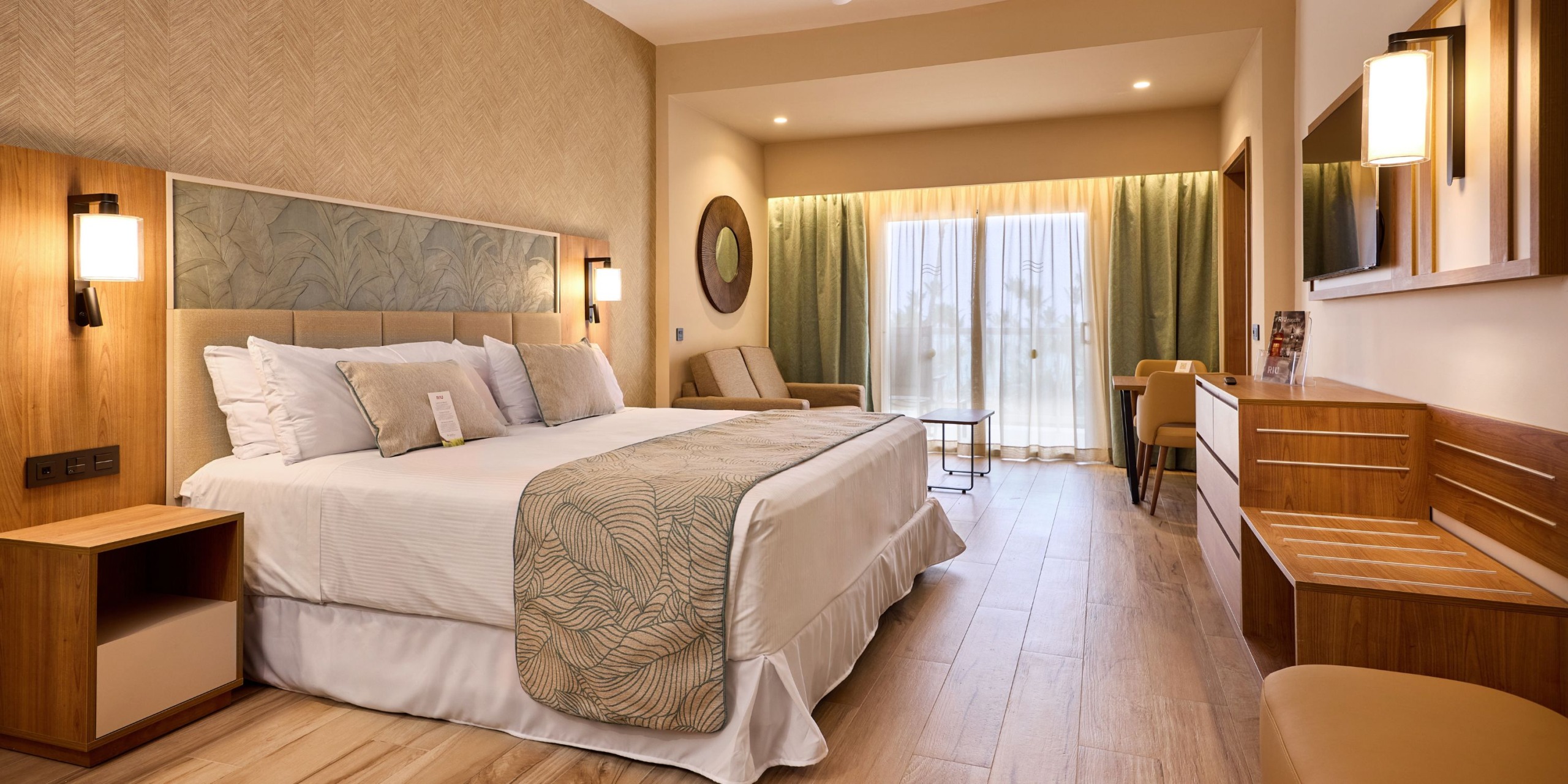 The Hotel Riu Palace Bavaro features a contemporary room equipped with a spacious bed, nightstands, a wooden desk, a round mirror, and a comfortable seating area. The room includes curtains that cover a large window and boasts flooring with a wood-like finish.