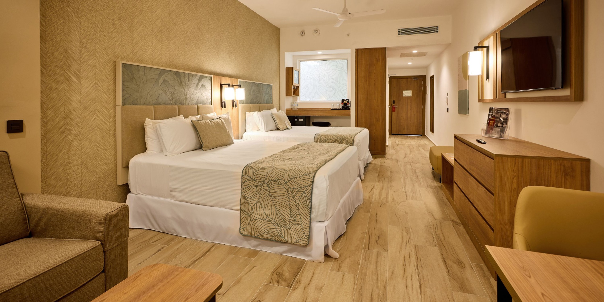 A room at Hotel Riu Palace Bavaro featuring two double beds, a cozy sofa, elegant wooden furniture, and a wall-mounted TV. The decor is light-colored, with a view that includes the bathroom entrance.